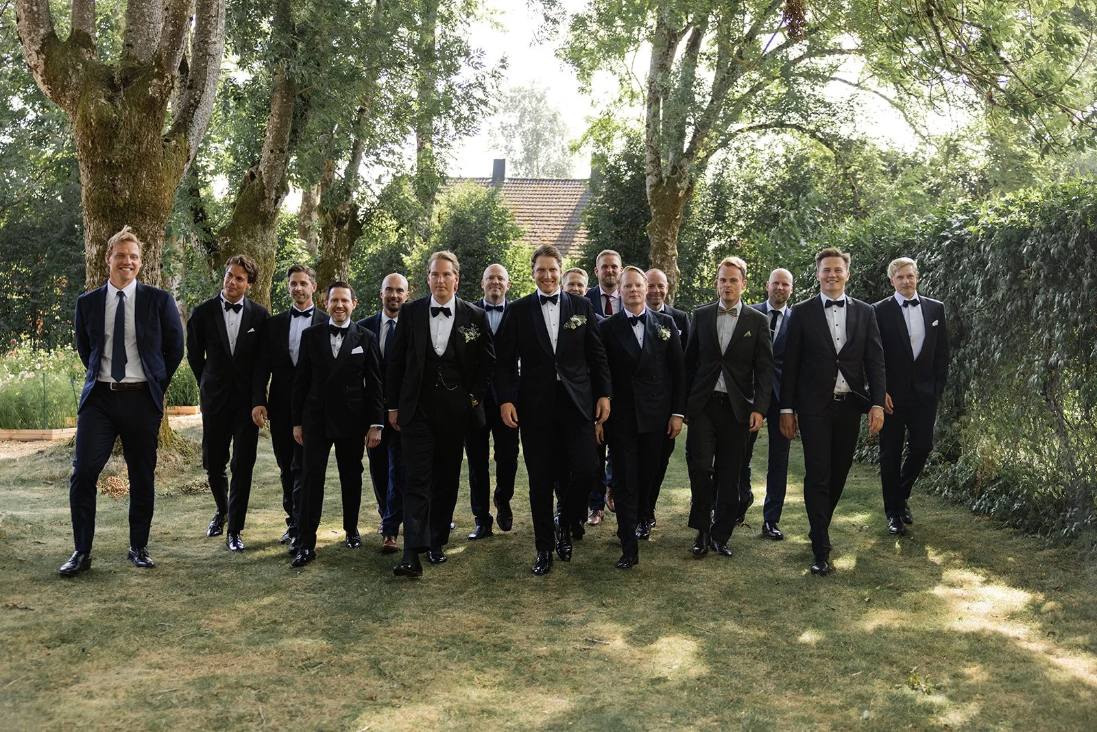 En gruppe menn kledd i svarte formelle dresser går gjennom en hage med store trær, under en solrik himmel.