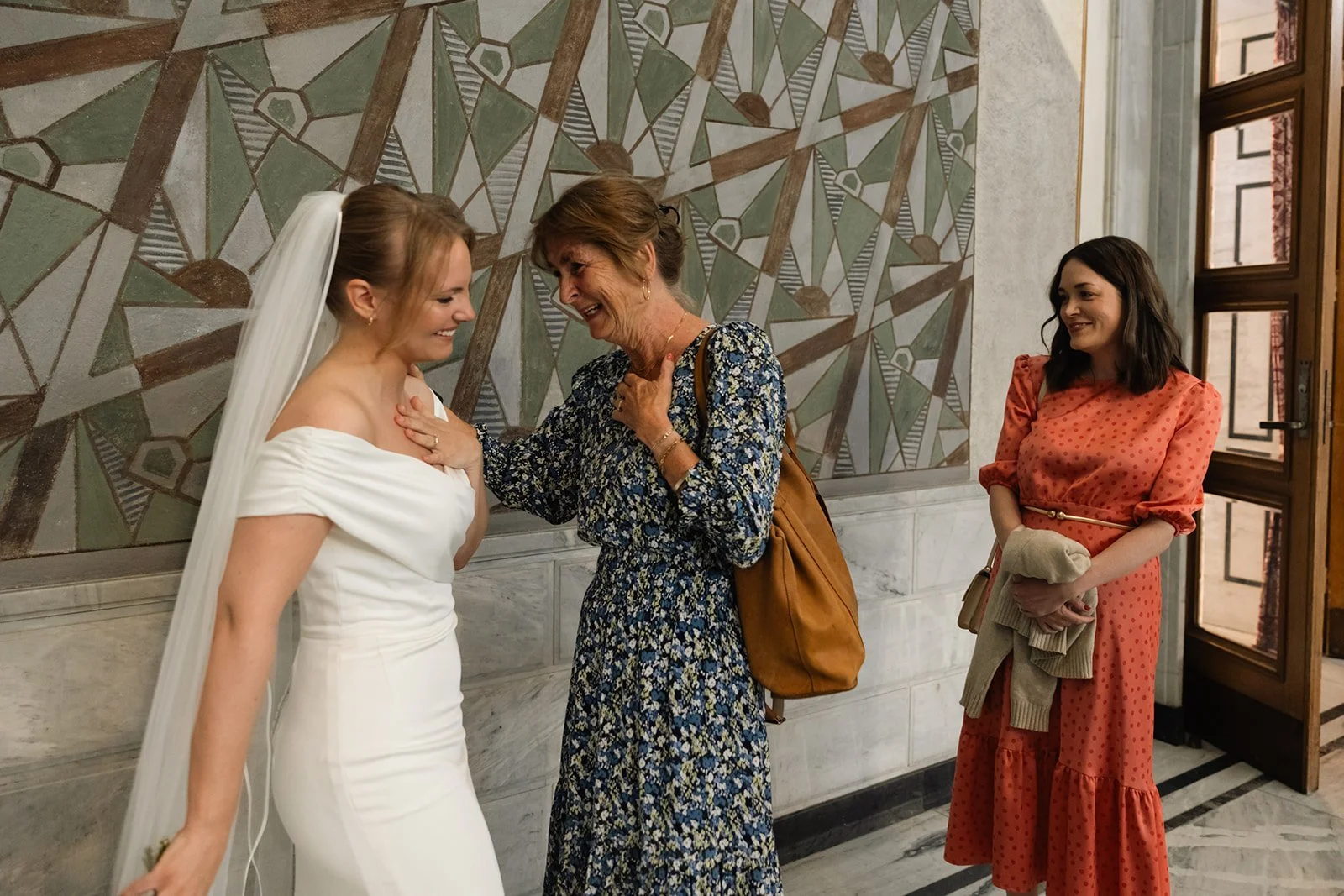 Tre kvinner i et rom, deriblant en brud i hvit kjole som ler, en eldre kvinne som smiler og papper, og en kvinne i oransj kjole som holder et skjerf og ser på.