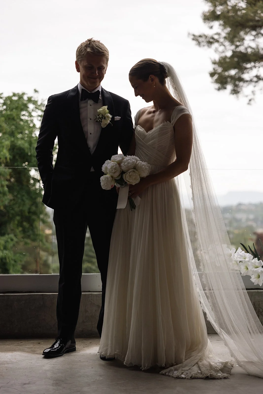 Et brudepar står sammen, kysses eller smiler, med brudekjole og dress, foran et utendørs landskap.