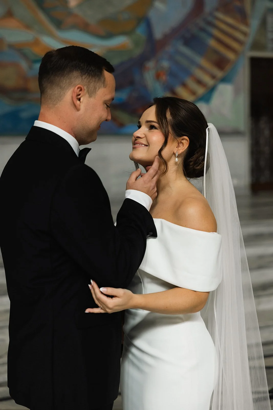 Et brudepar på bryllupsdagen, med mannen i sort frakk og kvinnen i hvit kjole og brudeblond, som ser på hverandre kjærlig i en innendørs setting.