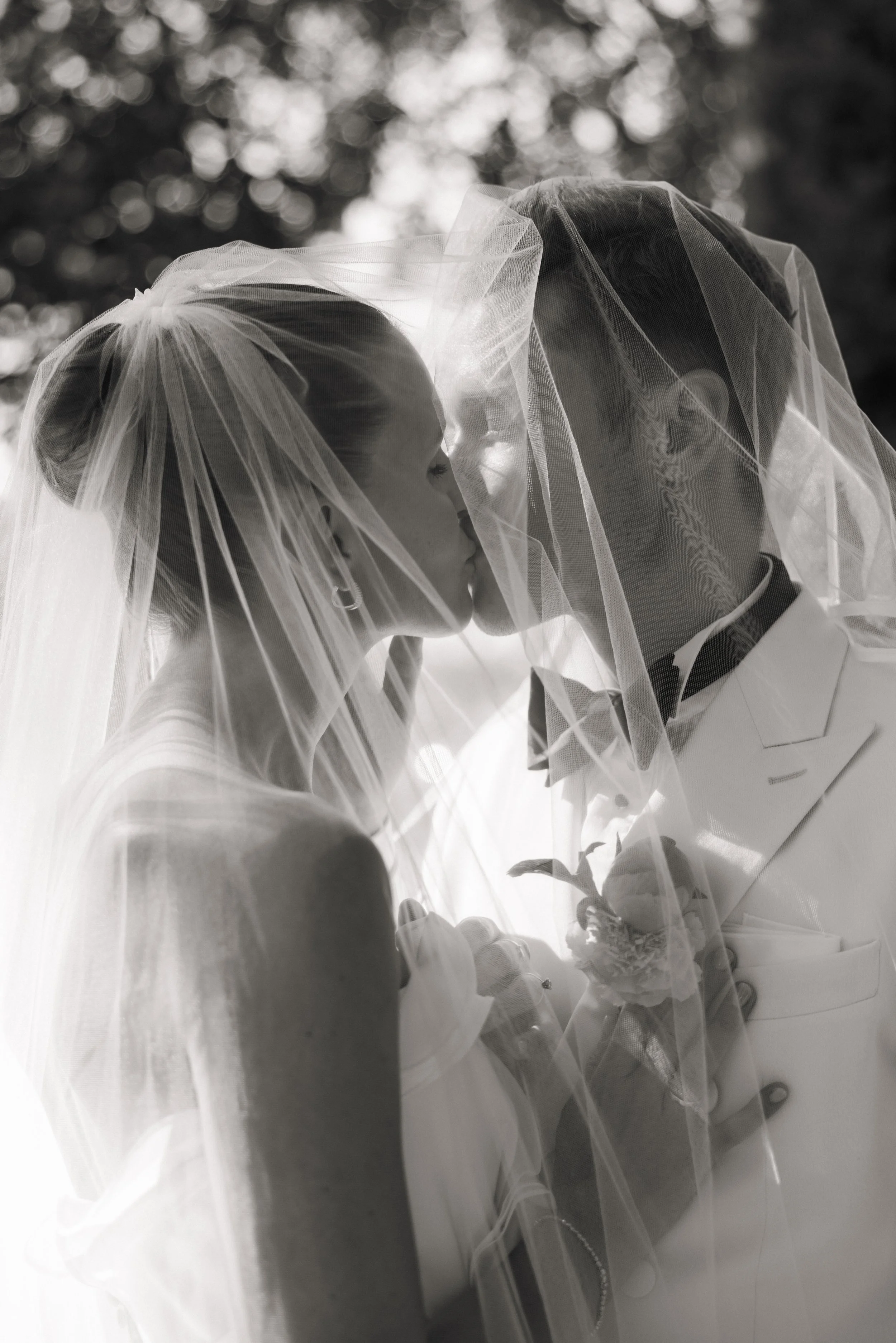 Et par på bryllup, der kysser under brudekjolenes slør, i svart-hvitt.