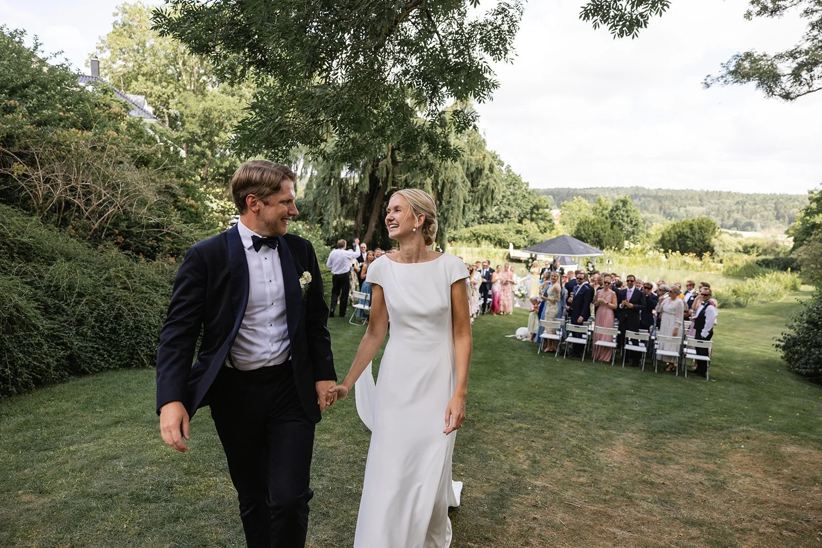En brud og en groom går hånd i hånd gjennom en hage under et bryllup, med gjester i bakgrunnen.