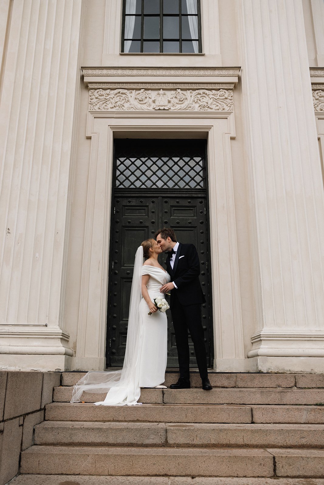 Nygift par kysser foran en stor bygning med svart dør og hvite søyler, bruden har lang hvit kjole og brudebukett, brudgommen har sort dress og slips.