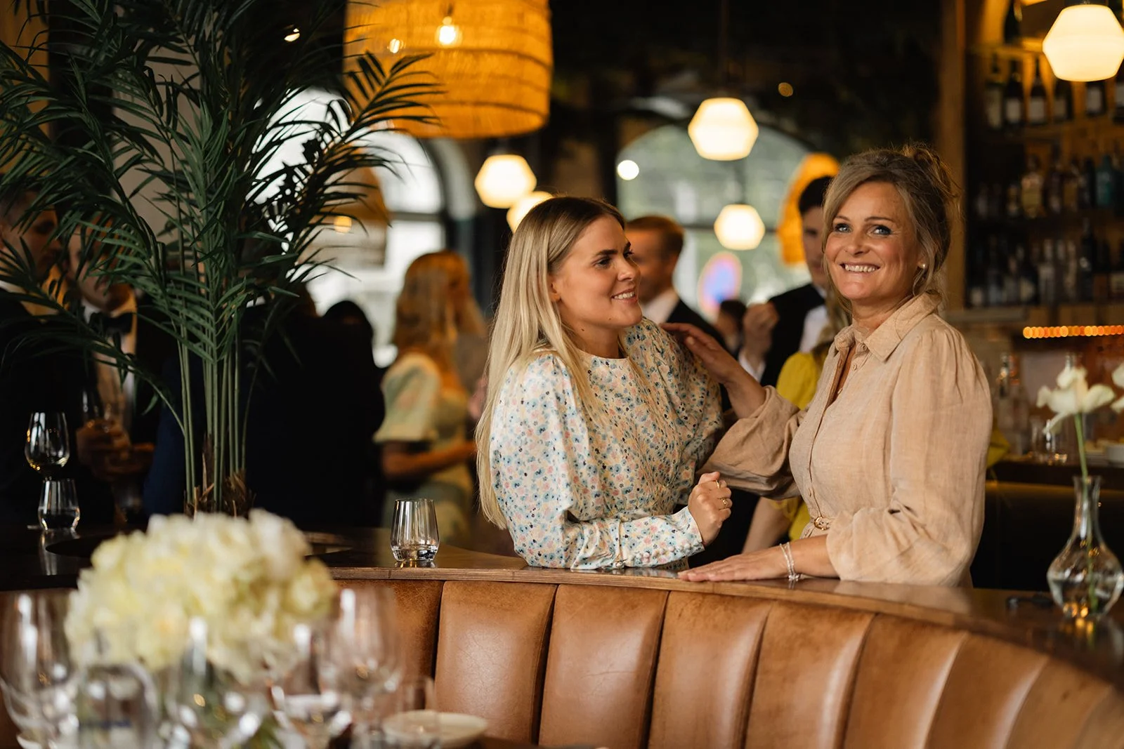 To kvinner ler og smiler mens de prater på en restaurant, med ølglass og blomsterdekorasjoner på bordet.