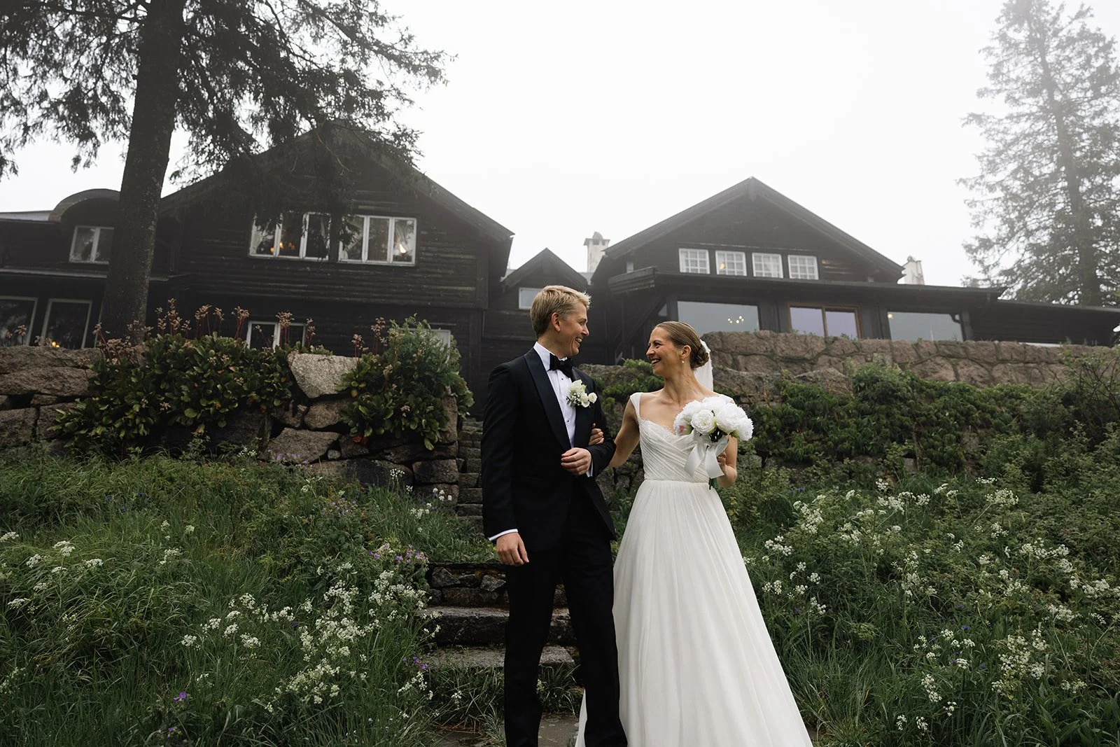 Et brudepar står sammen utenfor en svart trevilla, med grønne planter og blomster rundt seg, og smiler til hverandre.