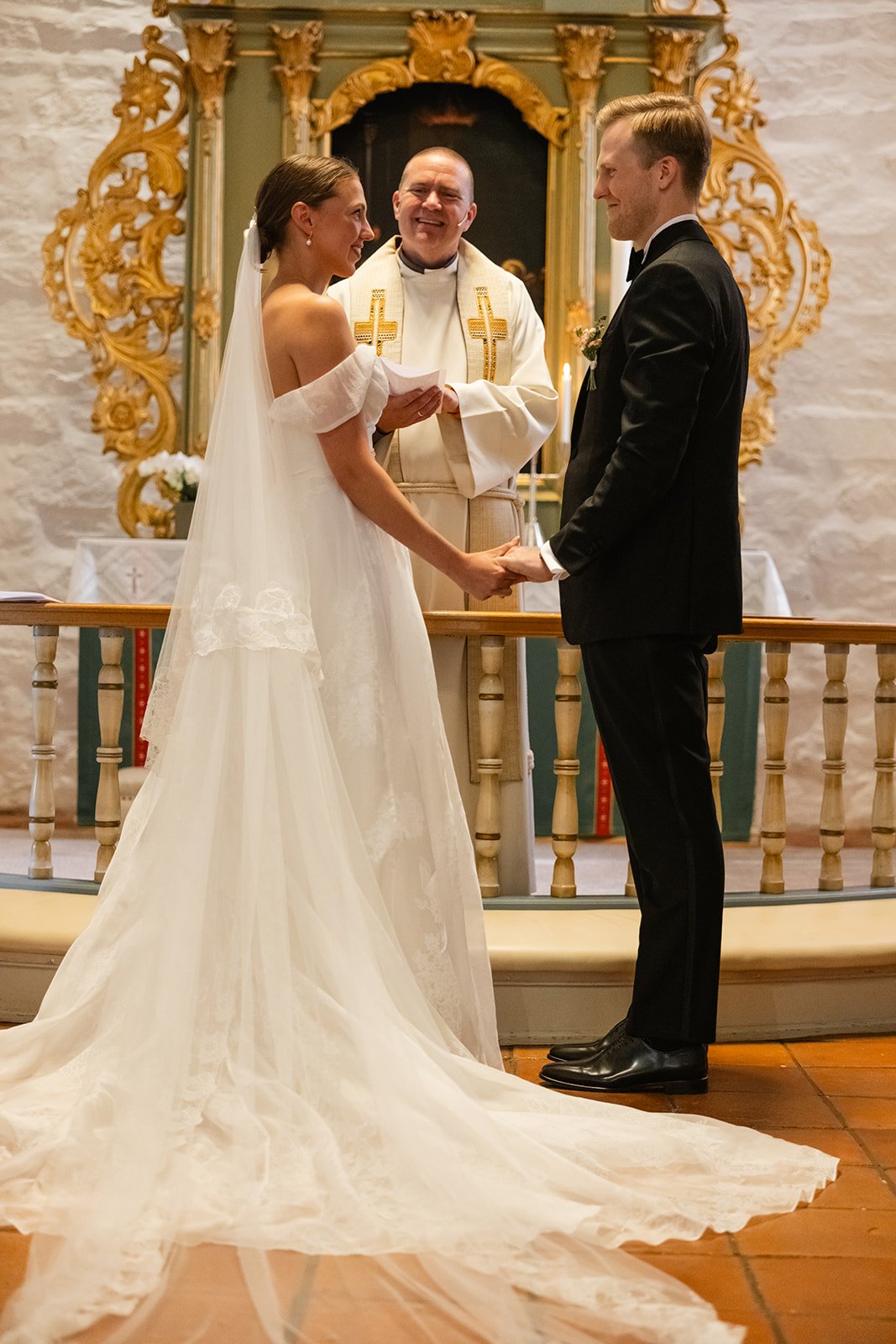 En brud og en brudgom holder hender under vielsen i kirken. Bruden har på seg en hvit kjole og brudgommen har på seg en svart dress. En prest står foran dem, og de er omgitt av dekorative gulldetaljer på alteret.