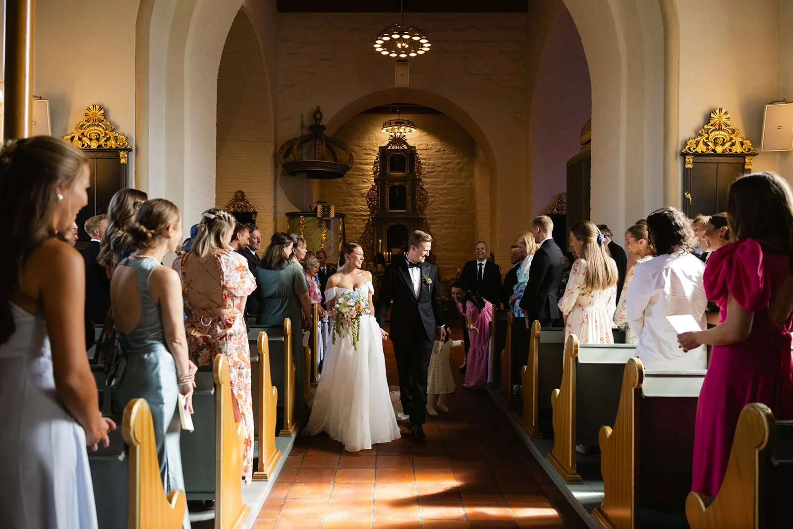 En bryllupsseremoni i en kirke med en nygift brud og brudgom som går ned midtgangen mens gjester ser på.