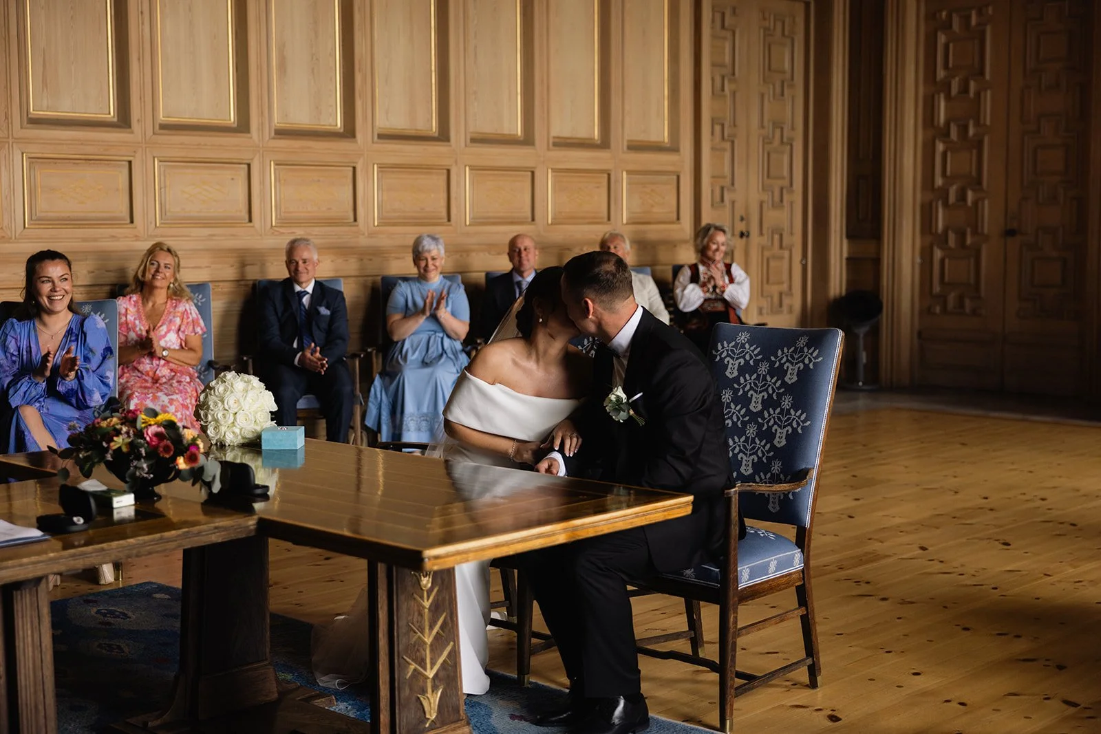 En brud og brudgom kysser hverandre under vielsen, omgitt av venner og familie i et rom med trevegger og gulv av tre.
