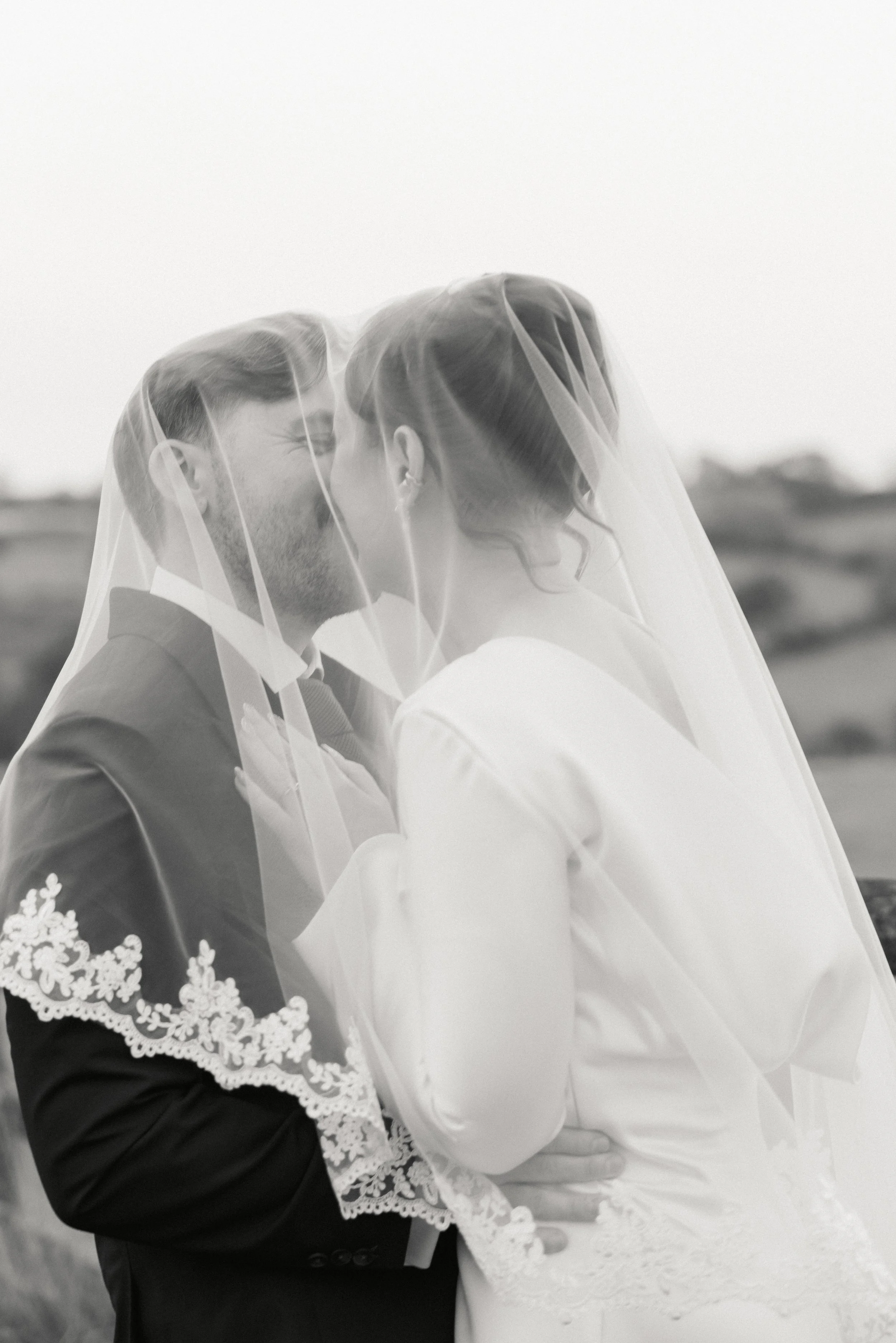 Et brudepar deler kyss under sløret i svart-hvitt fotografi.