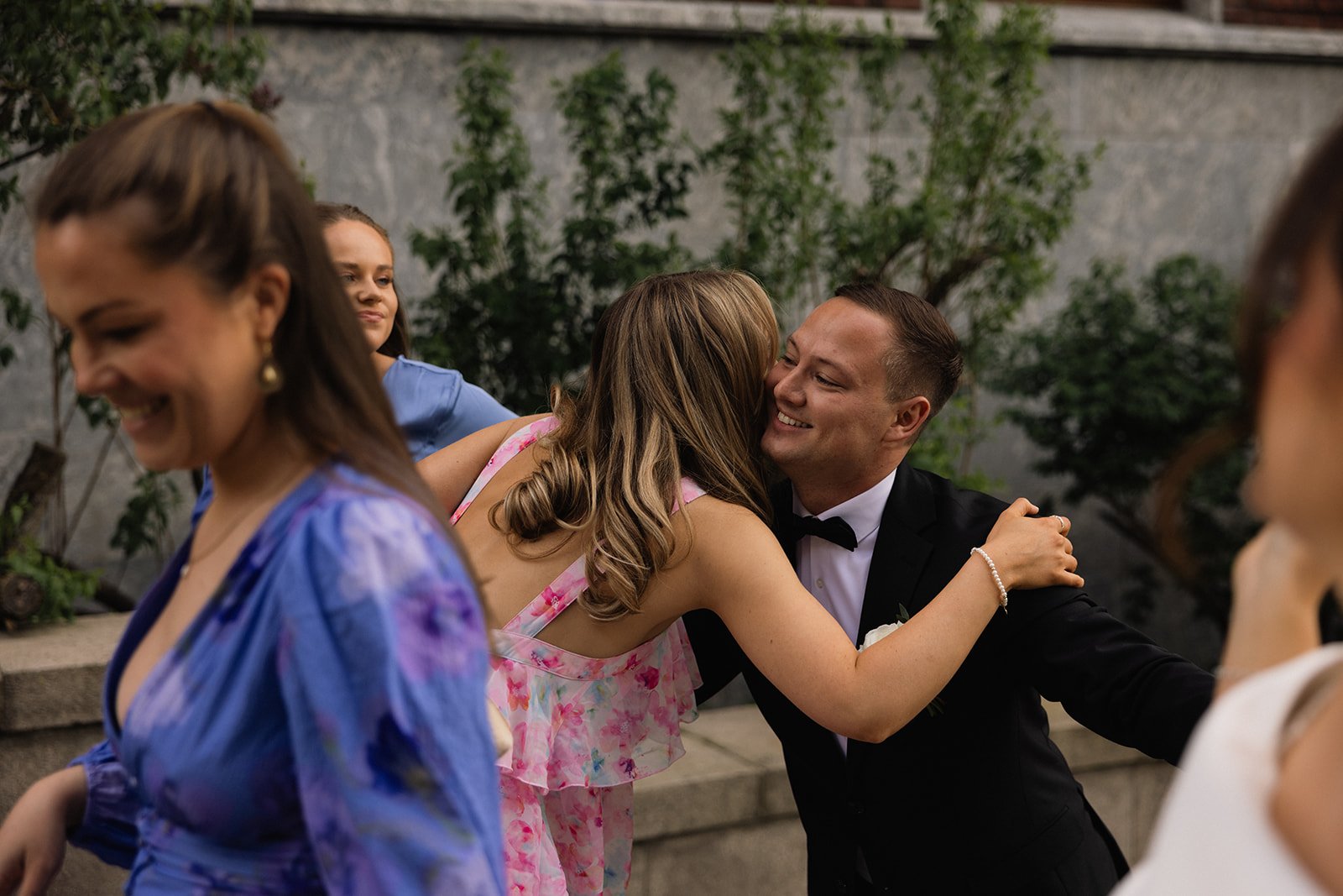 To personer som omfavner og gir en klem på en formell begivenhet, mens andre ser på, med blomstrende planter i bakgrunnen.