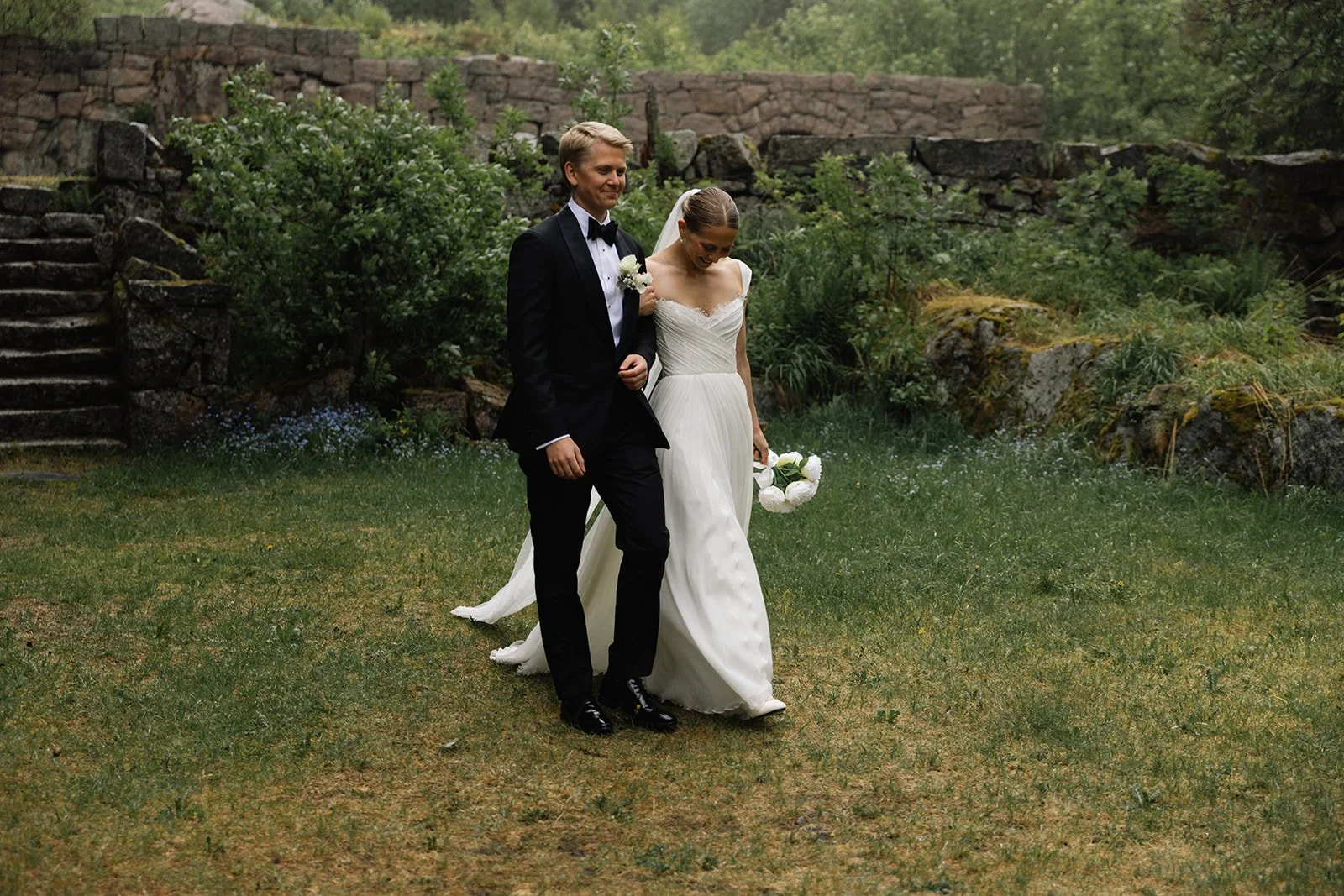 Et ektepar i bryllupsklær går sammen utendørs, omgitt av grønt landskap og gamle steintrapper.