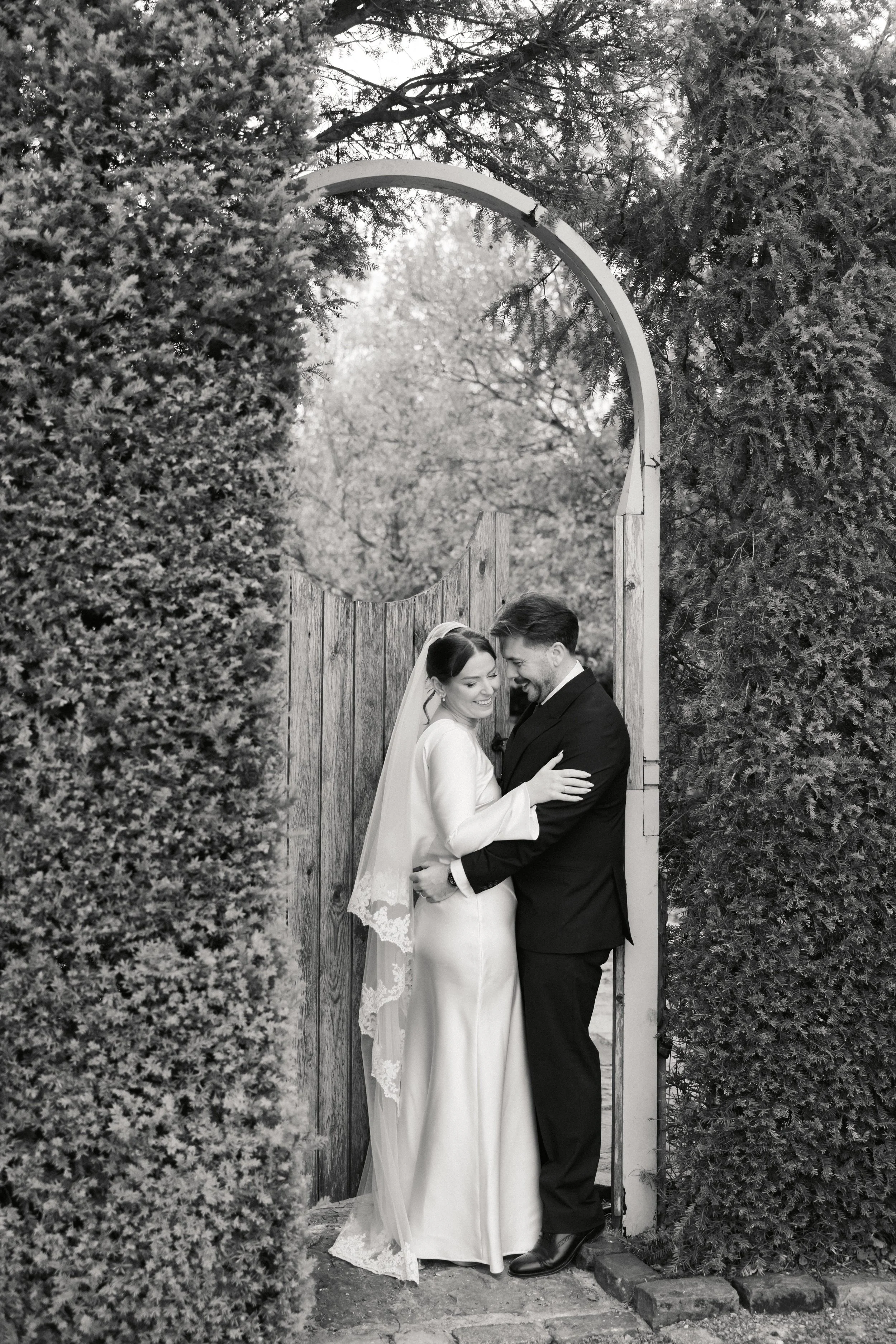 En brud og en brudgom som holder hverandre og smiler, fotografert foran en treport som er omringet av hekker, i svart og hvitt.
