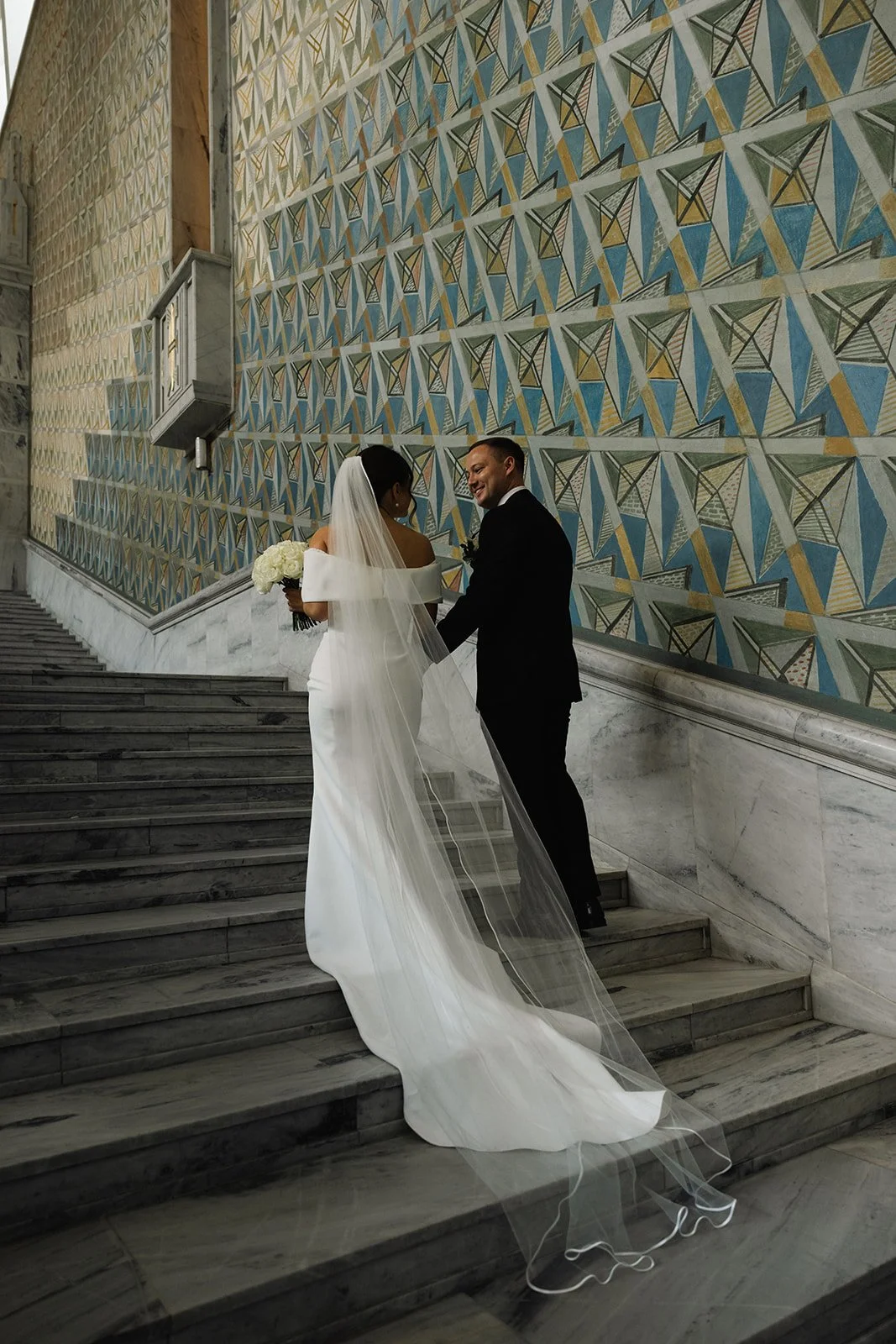Et bryllupsbilde av et par på trappen i en kirke med geometrisk Mosaikkvegg i bakgrunnen. Bruden har på seg en hvit kjole med lang, gjennomsiktig slør, og holder en blomsterbukett. Gommen har på seg en svart dress og hvit skjorte.