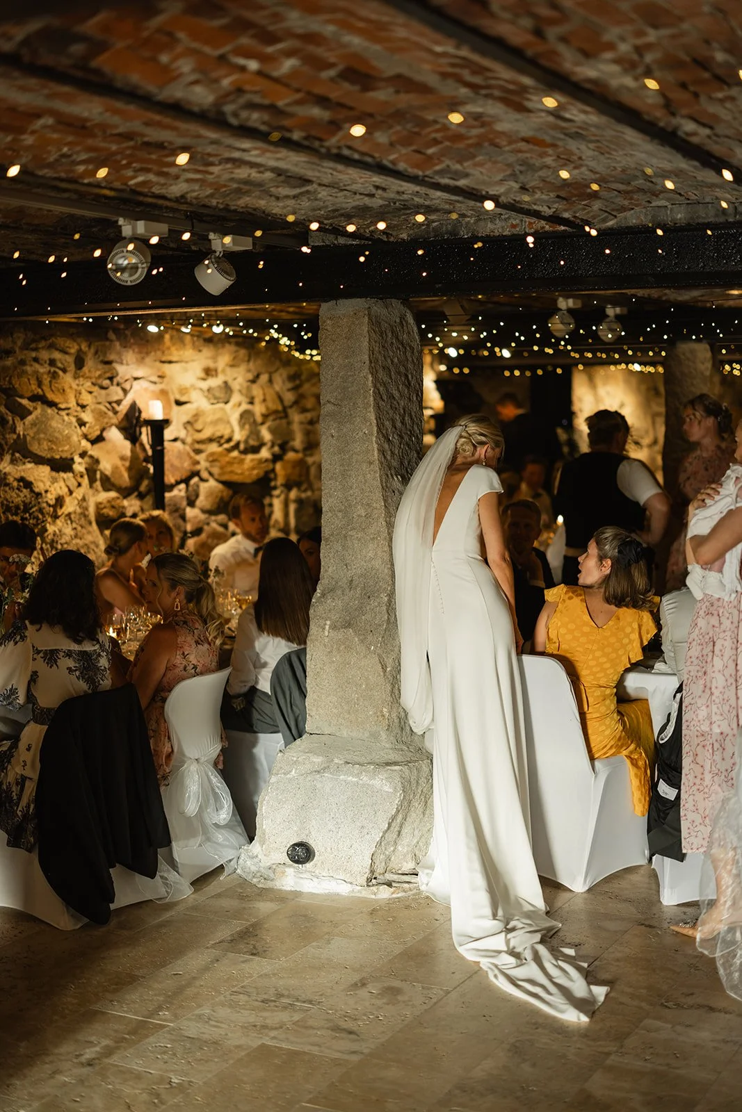 Kvinne i hvit brudekjole står i en låvetak med steinvegger, omgitt av bord med gjester ved bryllupsfest.
