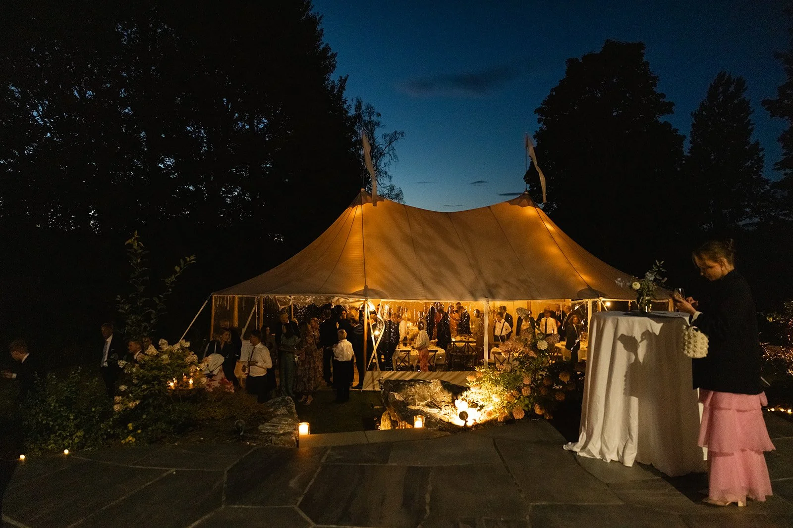 Et utendørs festligheter under en stor, opplyst gullfarget telt om kvelden, omgitt av trær.