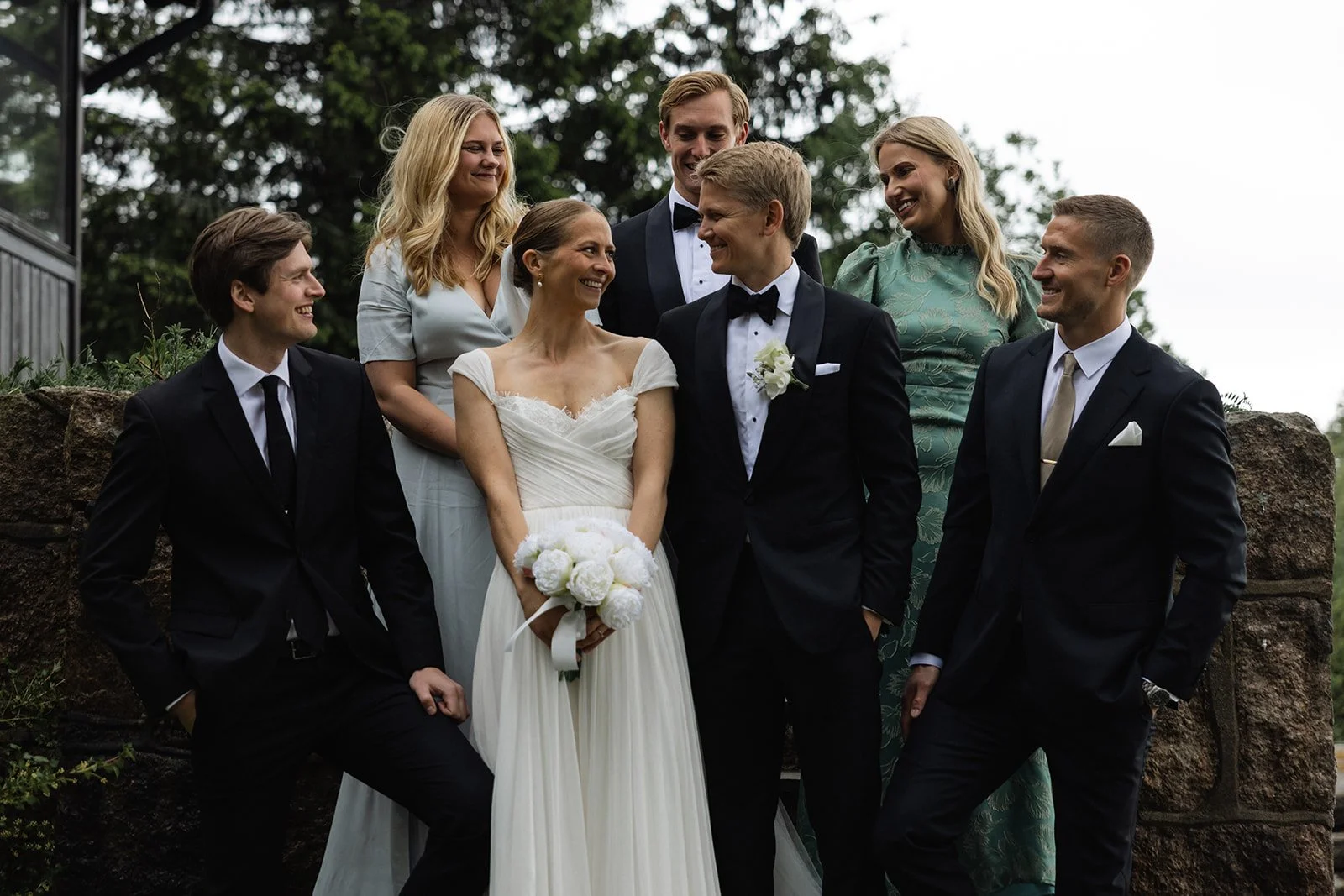 En gruppe mennesker i formelle klær, trolig på en bryllupsfeiring, står foran en steinmur utenfor. De smiler og ser på hverandre. En kvinne i midten holder en bukett hvite blomster.