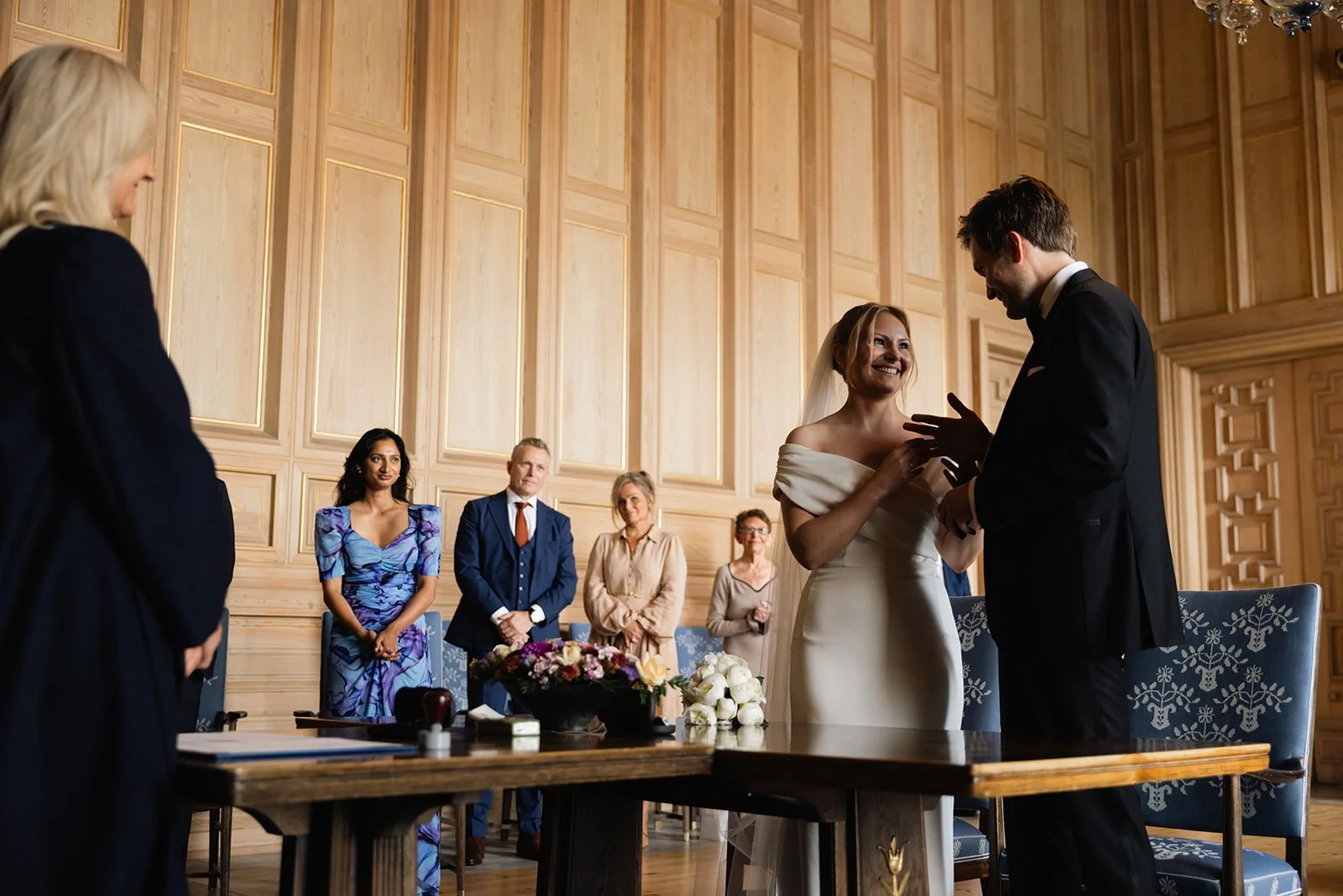 Et bryllupsceremoni med bruden og brudgommen som bytter løfter, omgitt av familie og venner i et rom med trevegger.