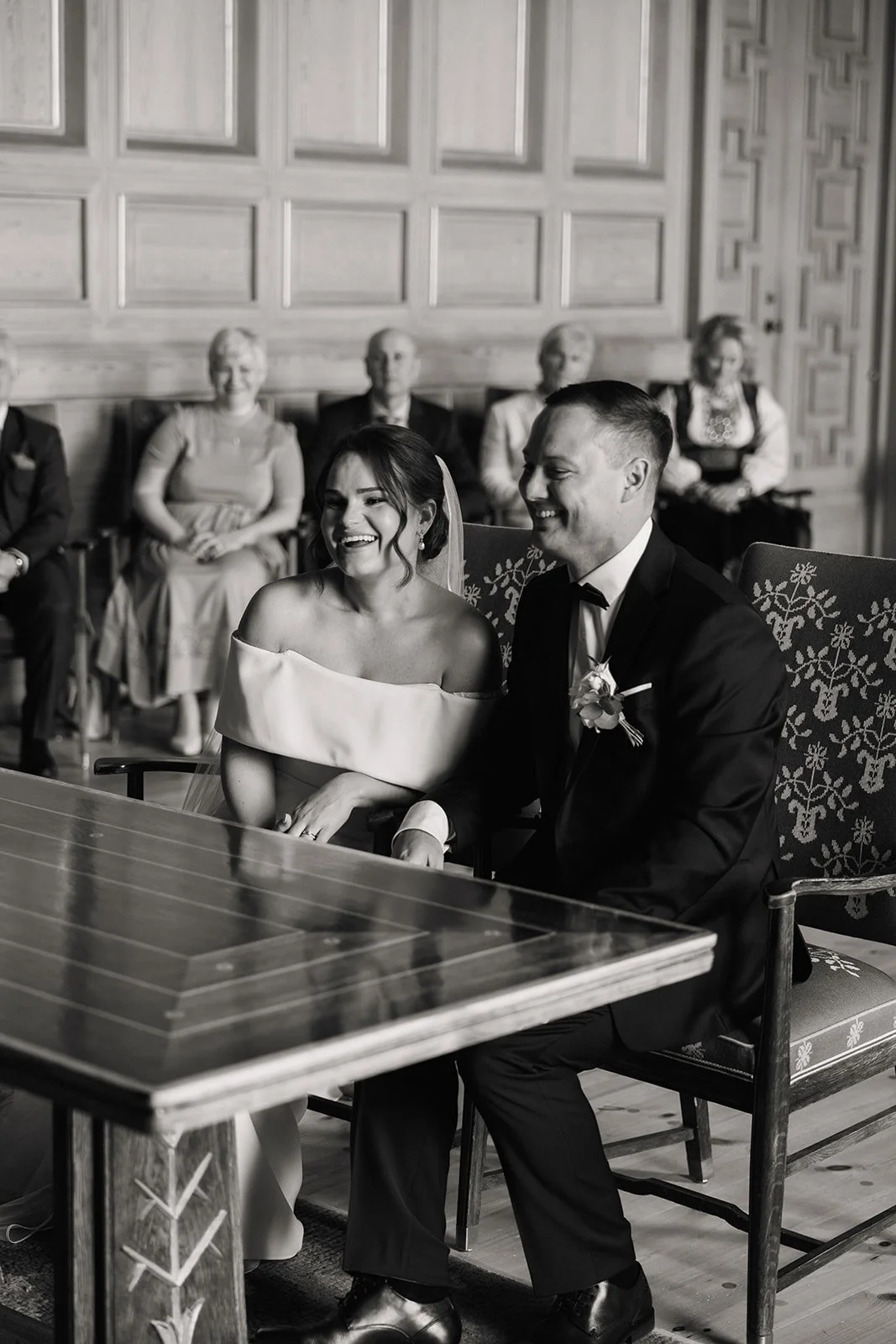 Et svart-hvitt bilde av et bryllup med en brud og en brudgom som sitter sammen foran et bord, omgitt av gjester i et rom med tresplanker på veggene.