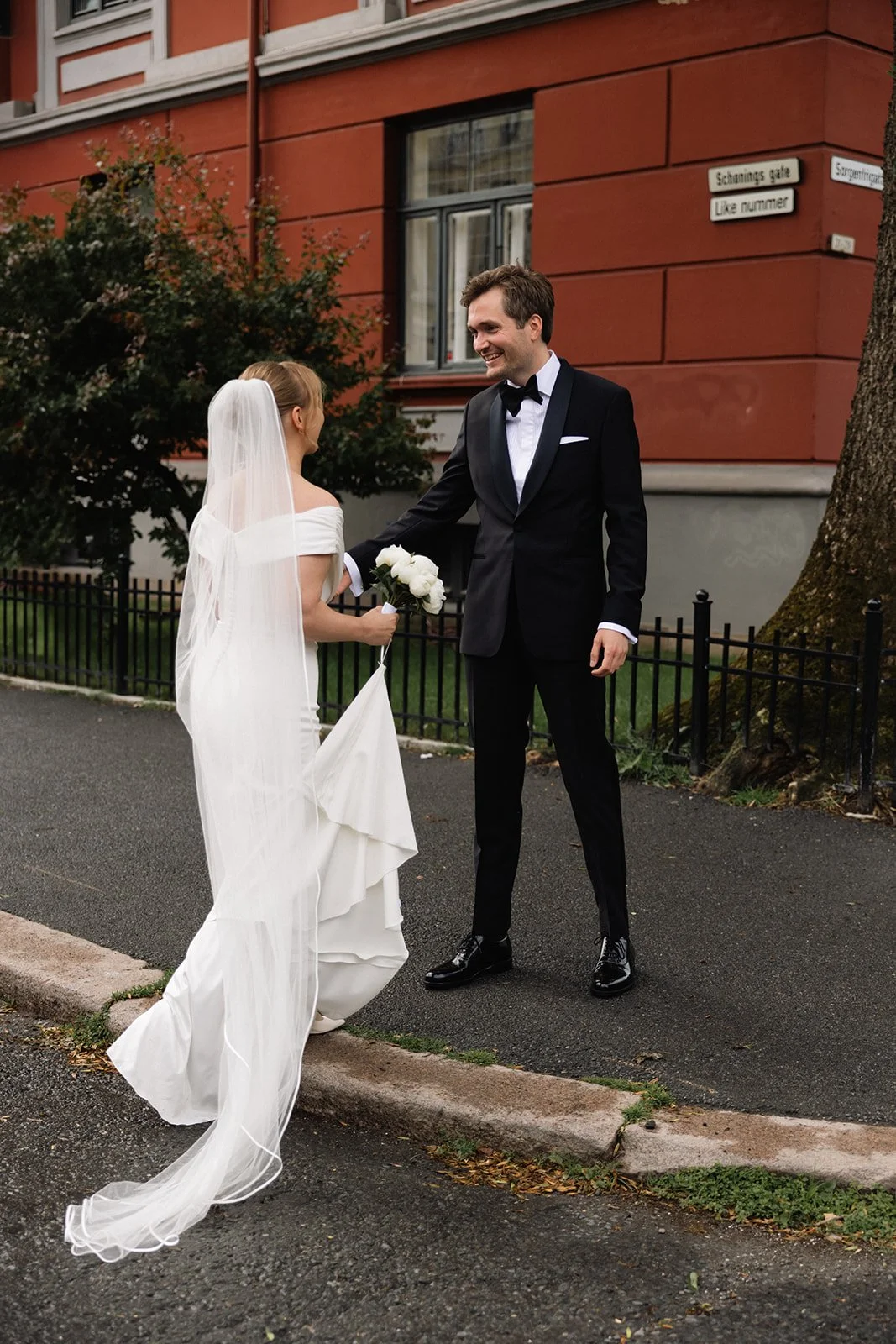 Et kledt brudepar står på gata, der kvinnen i hvit brudekjole og slør smiler og holder en blomsterbukett, mens mannen i svart smoking og sløyfe gir henne en hånd. Bygningsfasade i rød mur i bakgrunnen.