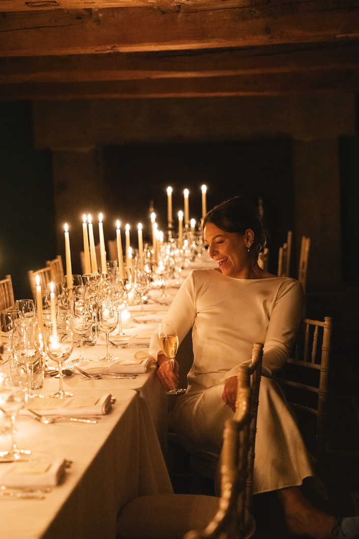 En kvinne i en elegant, hvit kjole sitter ved et langbord med tente lys, smiler og holder et glass champagne.