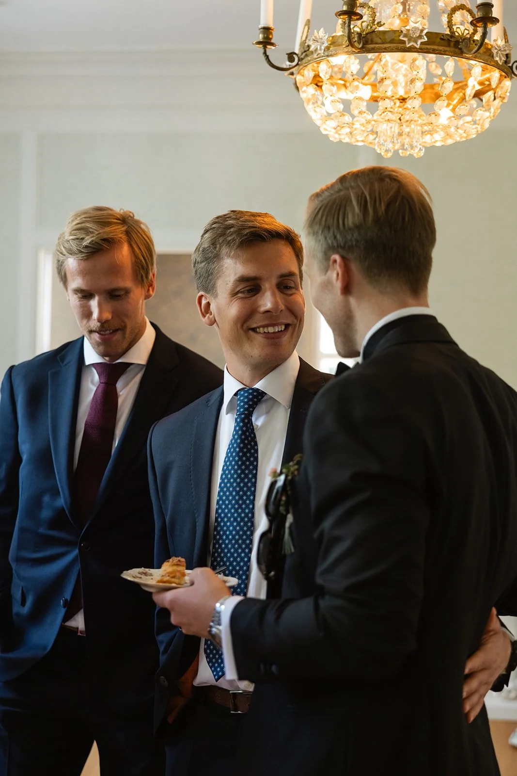 Tre menn i bunad og formelle klær snakker og ler under en lys krystallkronelysning.