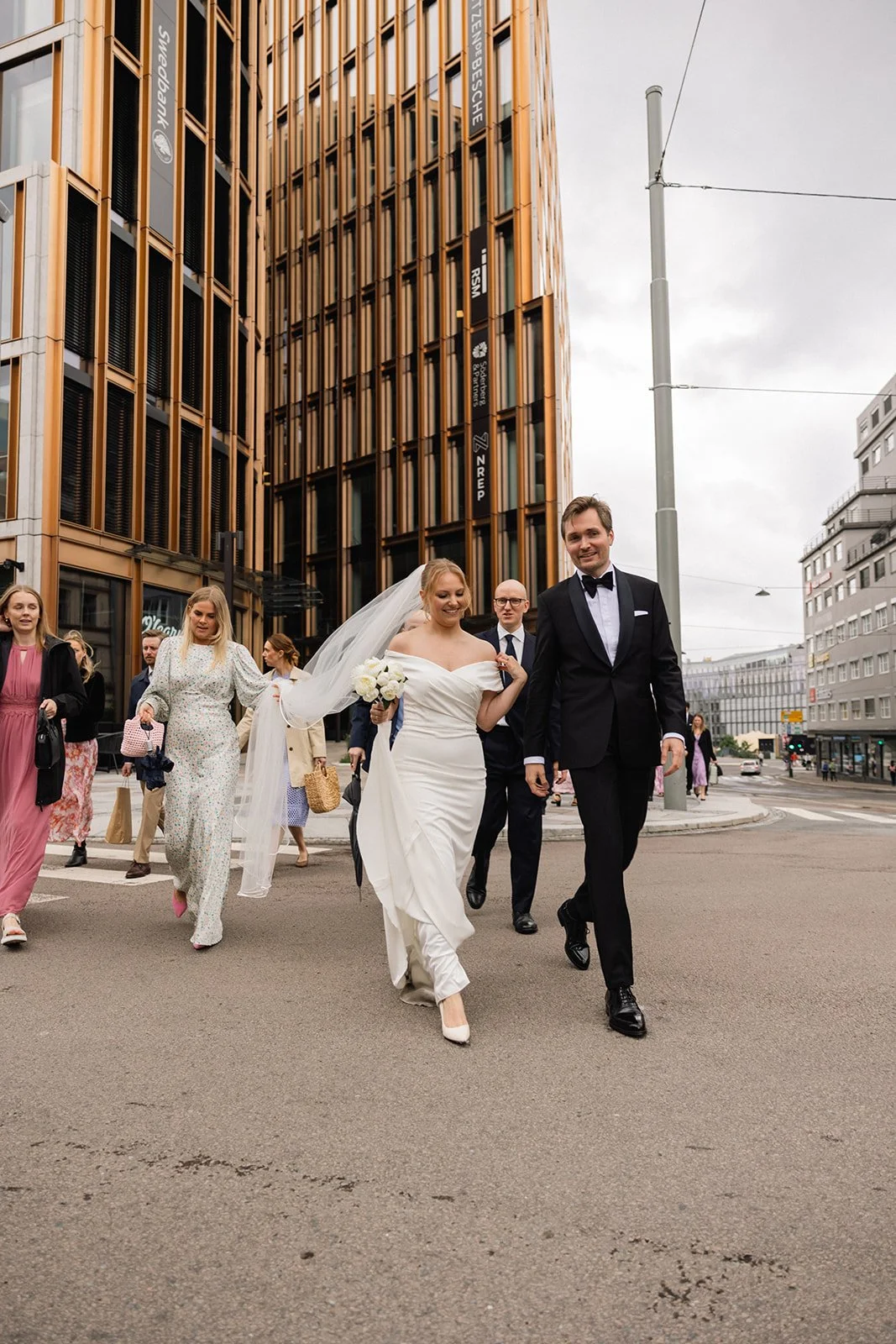 Et brudepar går lykkelig i bryllupsklede, omringet av venner, i en bygate med moderne bygninger og overveiende grått vær.