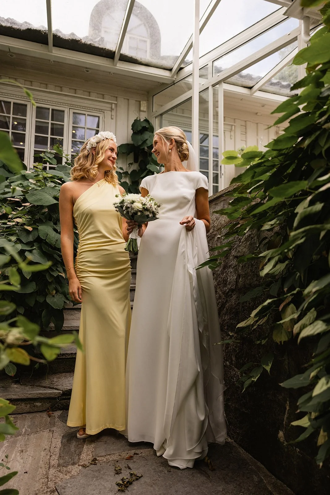 To kvinner står sammen i et glasshus den ene i en hvit brudekjole og den andre i en lys gul kjole, og de smiler til hverandre.