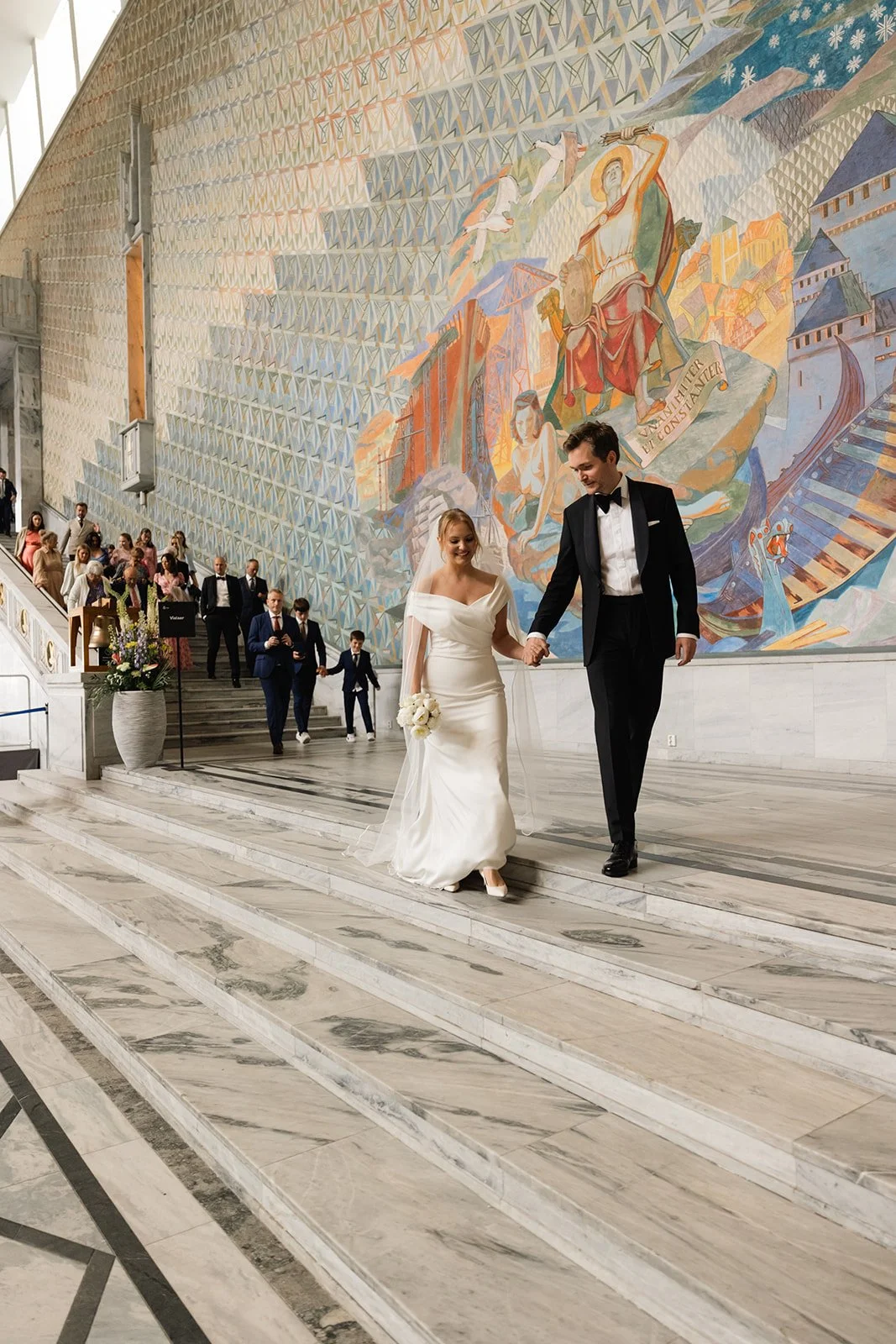 En brud og brudgommen går nedover trappen i en kirke, omgitt av gjester, med en stor fargerik veggmaleri i bakgrunnen.