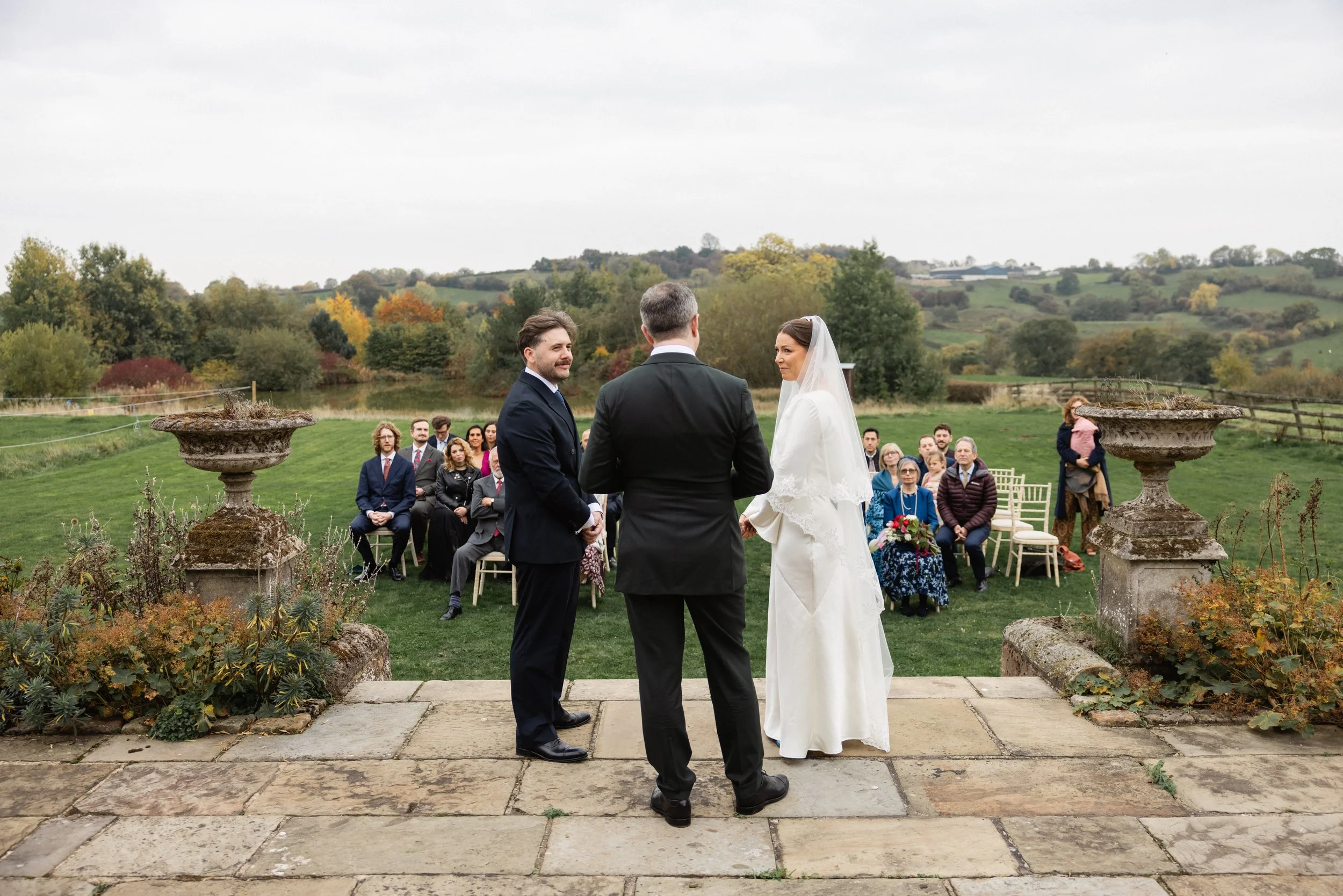 Et bryllup utendørs med brudeparet foran en liten gruppe gjester, omgitt av grønne åser og trær i høstfarger i bakgrunnen.