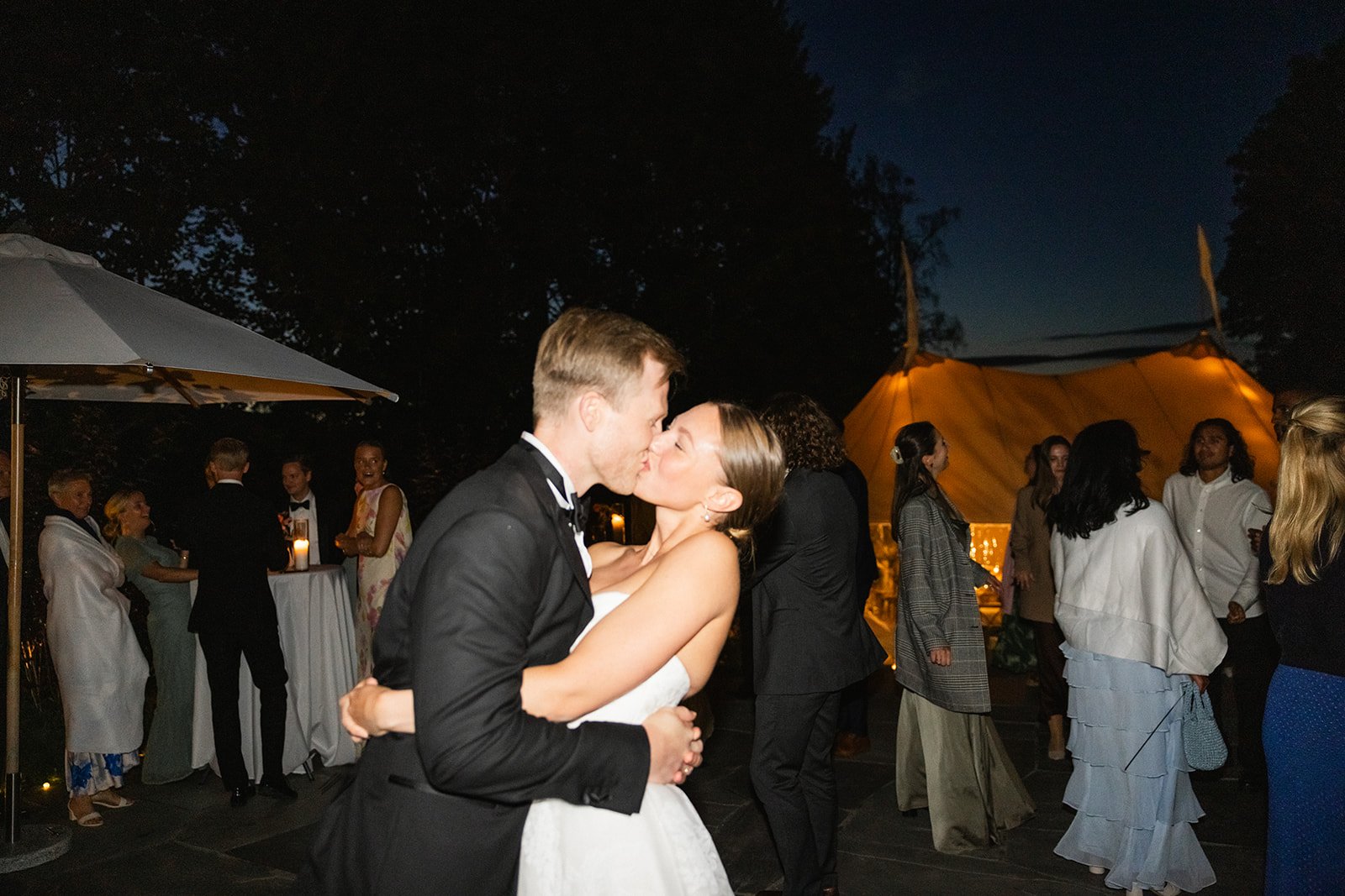 Et kveldsbilde av et bryllup med et par som kysser, omgitt av gjester som snakker og danser utendørs under en stor, opplyst telt.