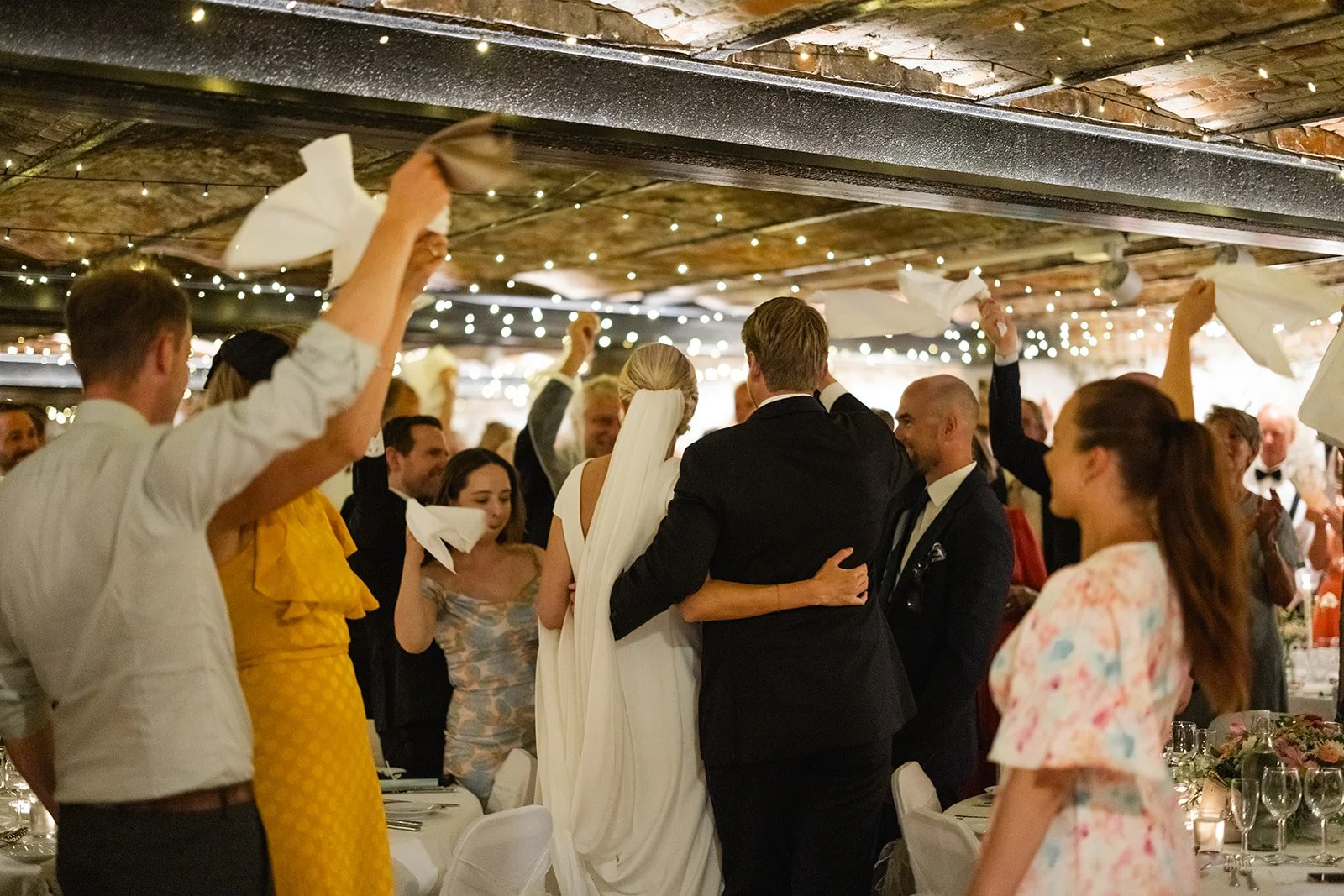 Brudepar og gjester feirer i en festlokale med dekorasjoner og lys, mange hever lommetørklær i en rørende stund.