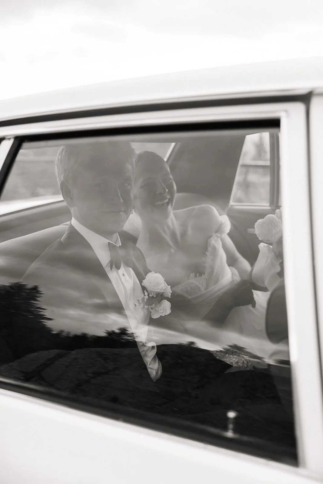 To personer i brudekjole og dress sitter i en bil med kommunikasjon og smiler, sett gjennom et bilvindu.