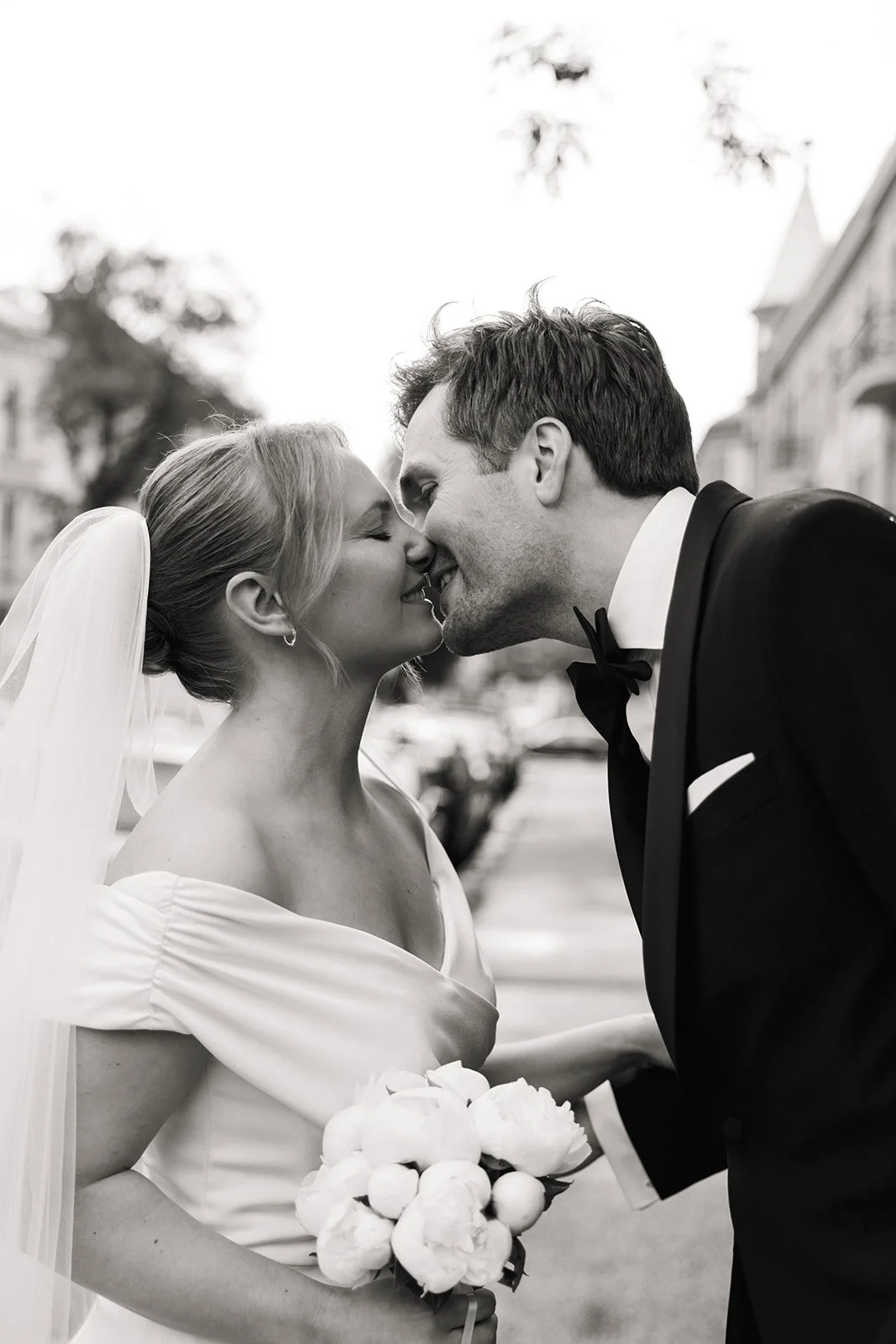 Nærbilde av et nygift par som kysser med nesene inn mot hverandre, den kvinnelige bruden holder en bukett blomster, de er kledd i bryllupsantrekk, og bakgrunnen viser en gate med parkerte biler og trær.