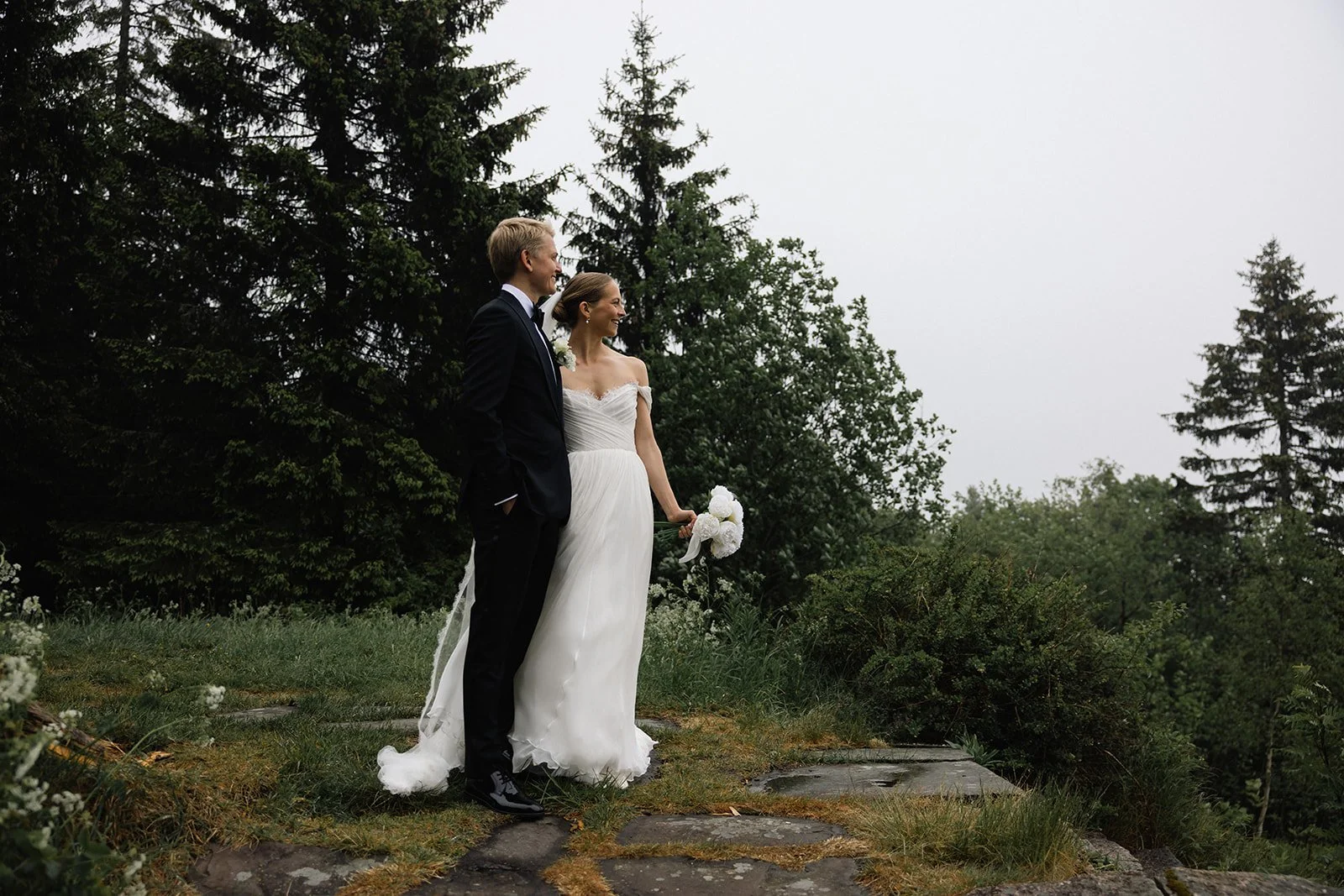 Et brudepar står på en steinlagt sti med trær i bakgrunnen, kledt i bryllupstøy, med kvinnen i en hvit kjole som holder en bukett hvite blomster.