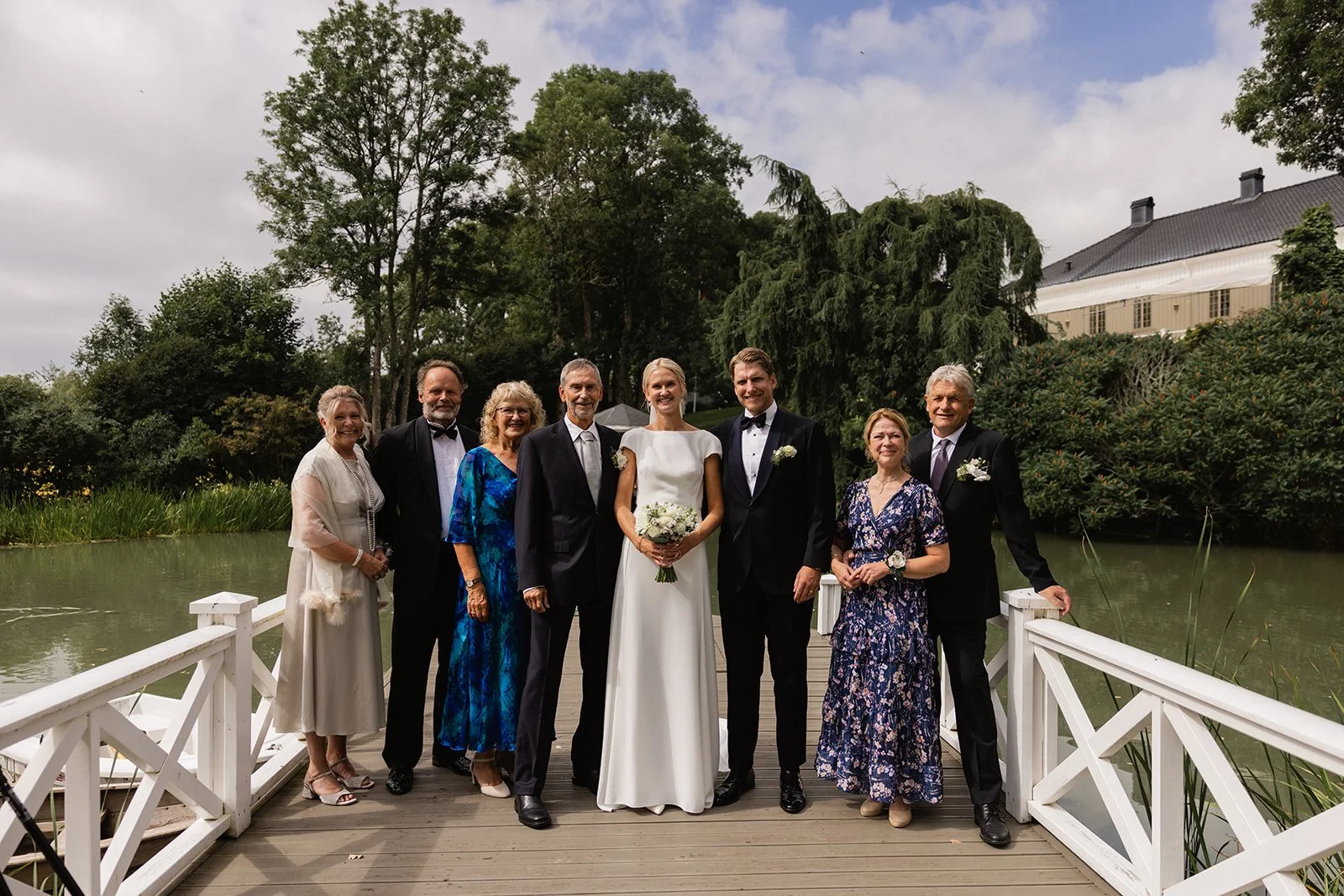 Bryllupssans på en bryllupsdato med ni personer på en brygge ved vann, omgitt av grønne trær og en bygning i bakgrunnen.