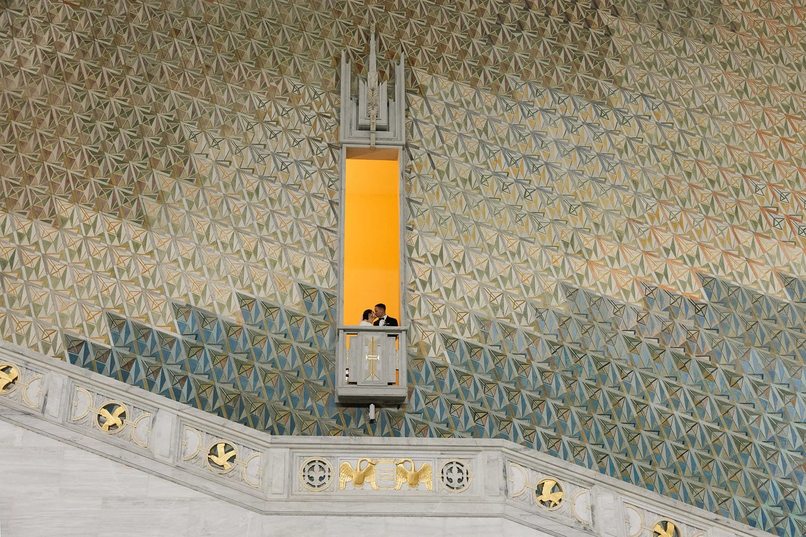 Et par i bryllupsantrekk står på en liten balkong innenfor et geometrisk dekorert vegg i en kirke.