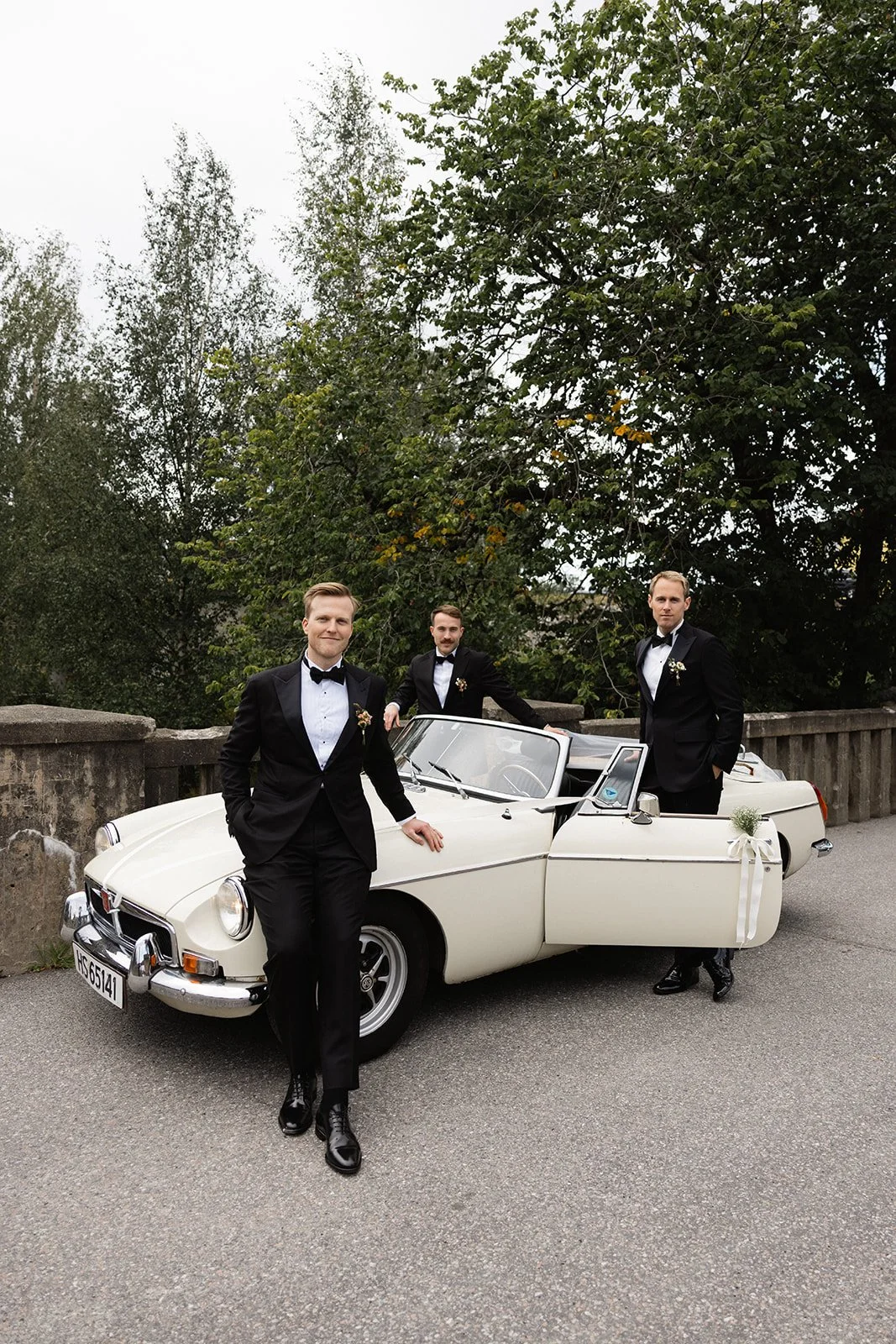 Tre menn i svarte smoking står ved en hvit vintage åkestol, med en stor tre og grønt løvtrær i bakgrunnen.