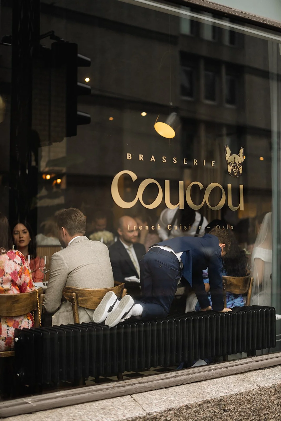 Utsikt gjennom en restaurantvindu med folk som sitter og spiser, og en gutt som klatrer på radiatoren. Restaurantens navn er Coucou, som serverer fransk mat i Oslo.