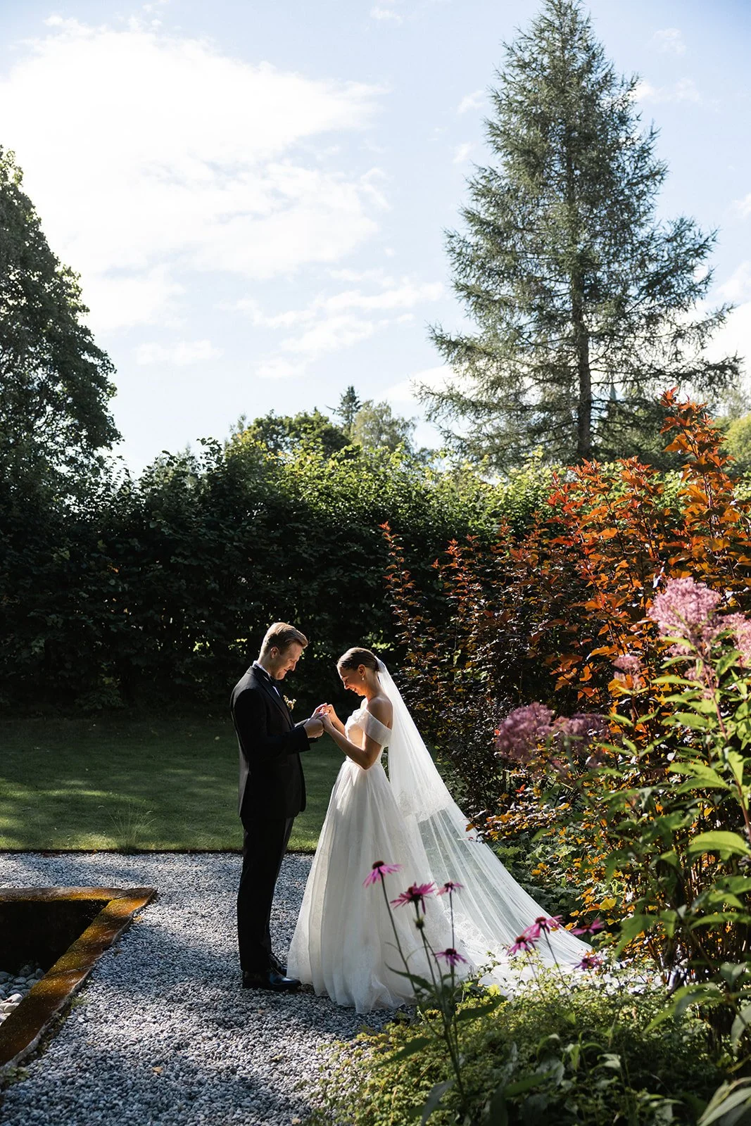 Et brudepar står i en hage, holder hender og ser på hverandre, omgitt av grønne planter og blomster, med en stor gran bakgrunn, under en klar himmel.