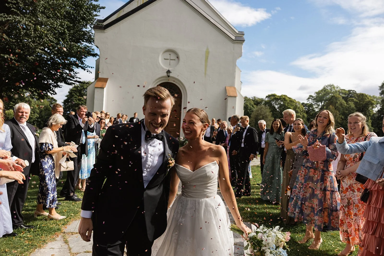 Nykgoende brudepar går ut av en hvit kirke mens gjester kaster blomster og korn.