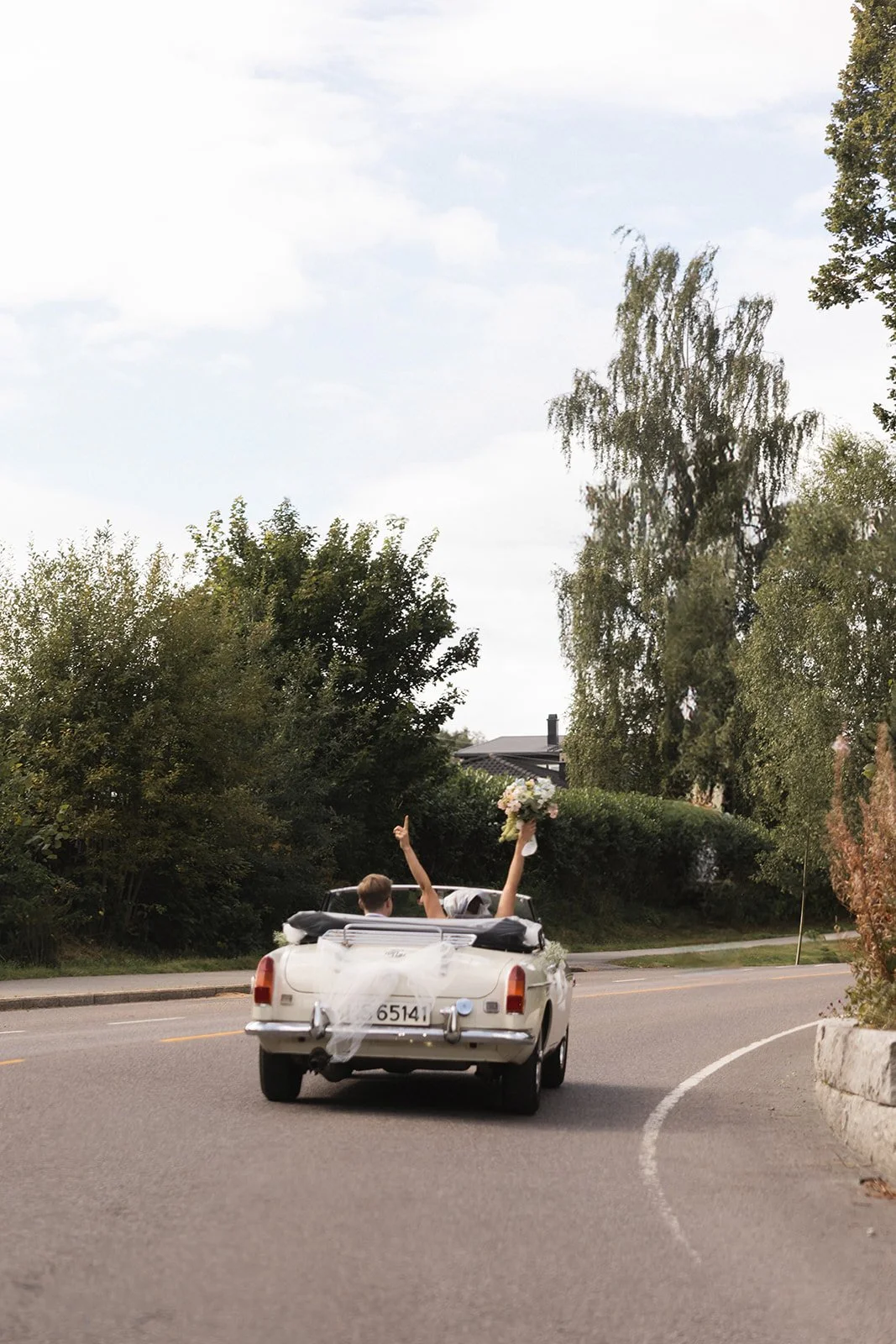 Et bruddpar kjører i en sving med en brudepike som holder en blomsterbukett, vinkende med den ene armen opp. Bilen er dekorert med hvitt stoff og har norske nummerplater.