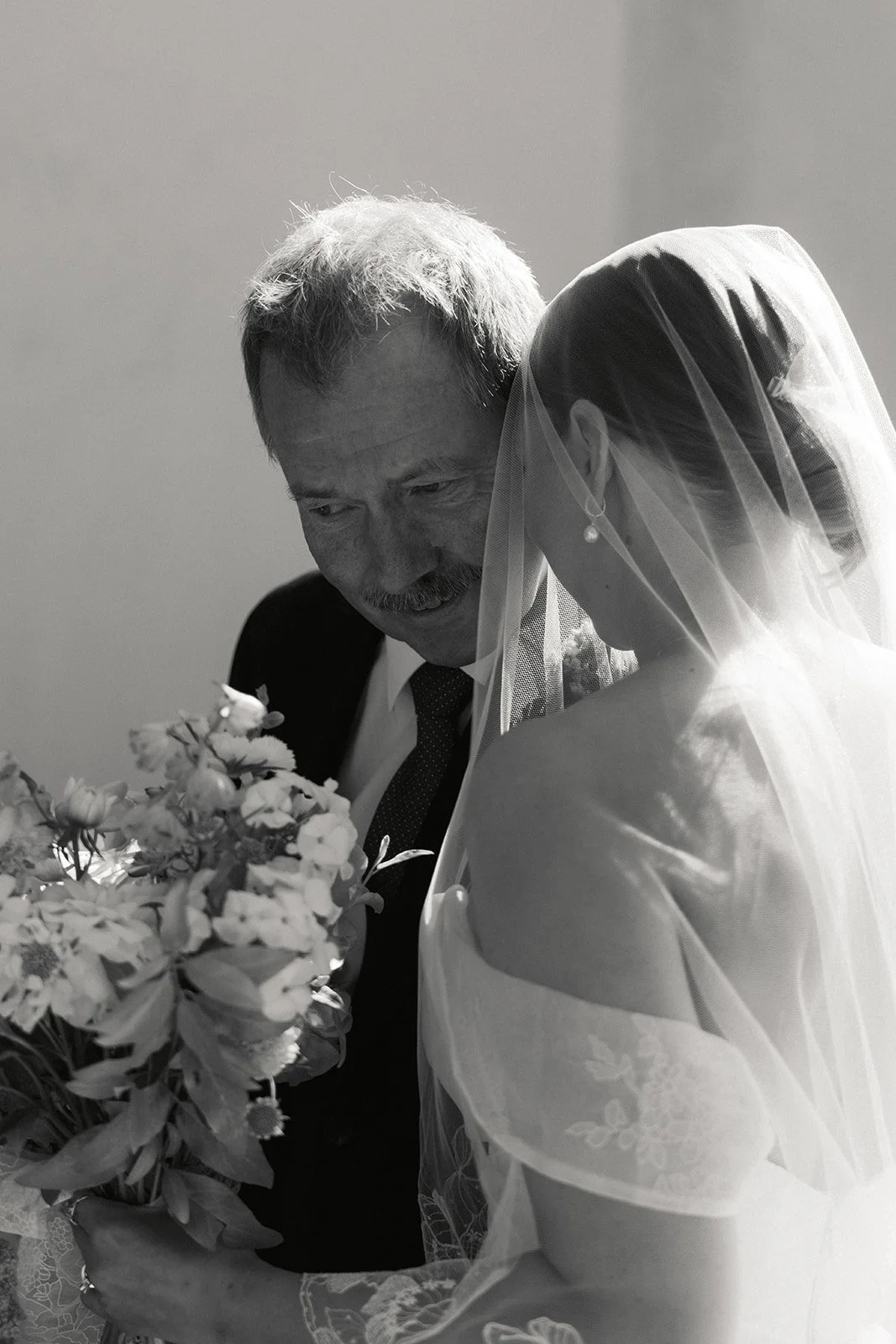 Et eldre manns ansikt smiler mens han ser på en kvinne som har på seg en brudekjole og et brudeslør, og de står foran en blomsterbukett.