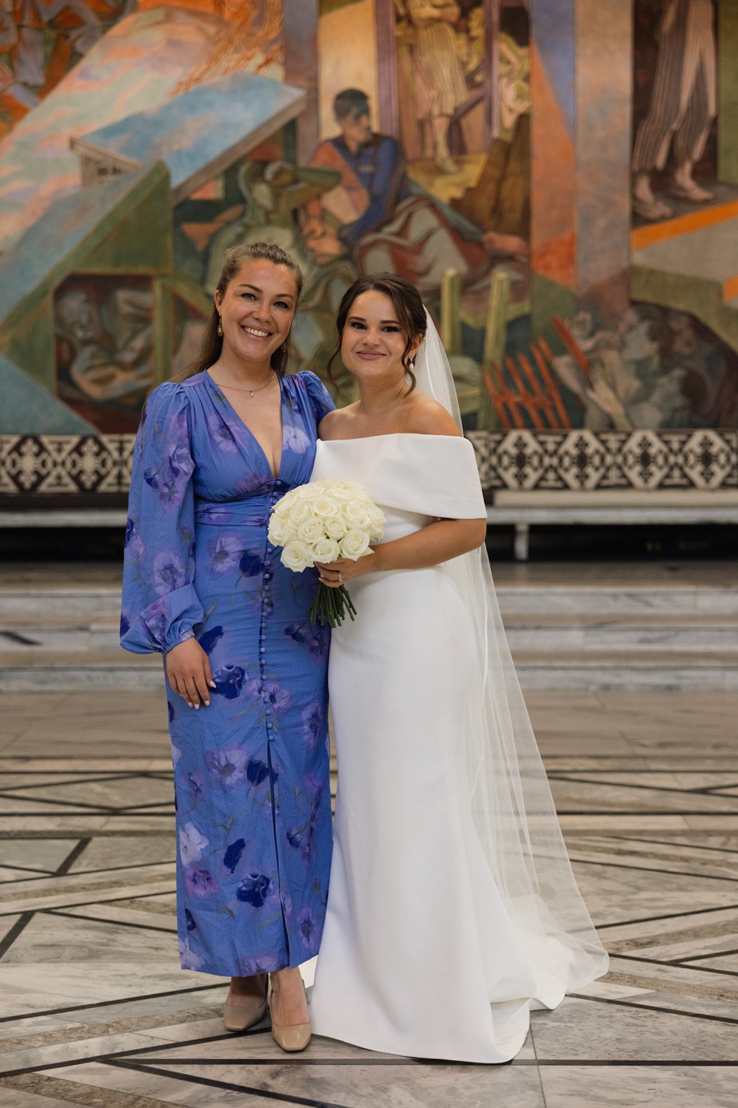 To kvinner står sammen i et kirkerom, den ene kledd i en hvit bryllupskjole med blomsterbukett, den andre i en blå kjole med blomsterprint.