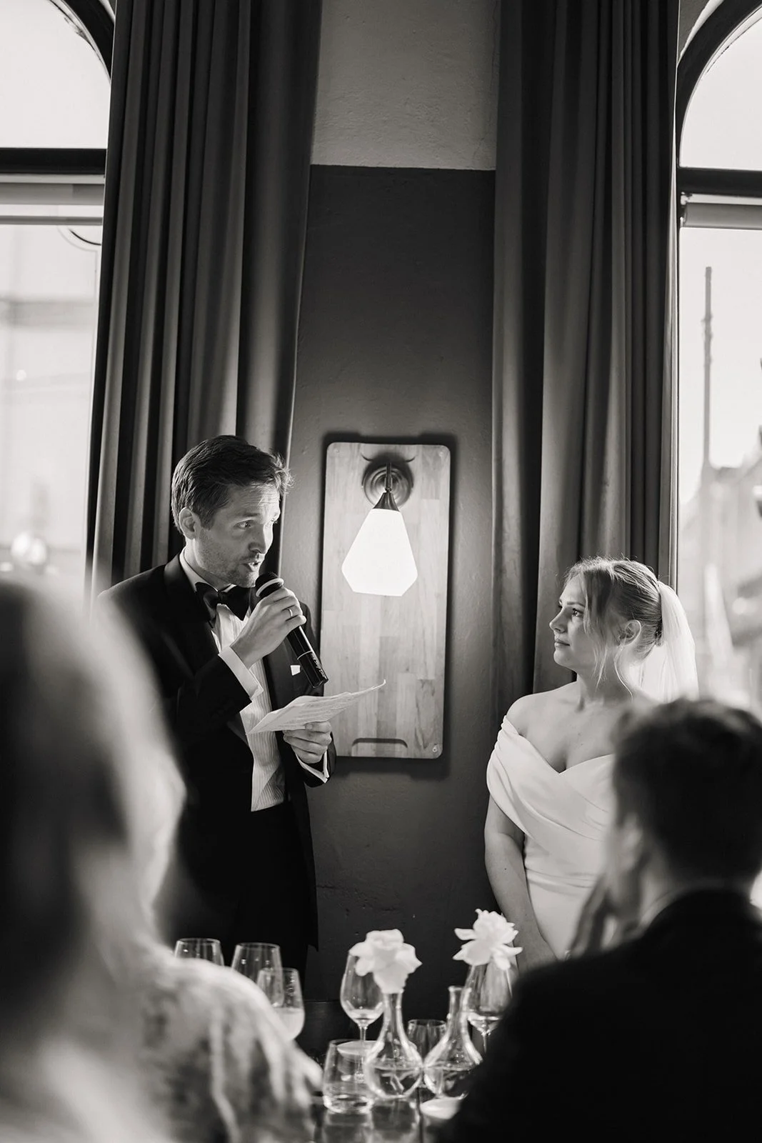 En tale holder for en brud og en brudgom under et bryllup. Brudgommen leser sannsynligvis et løfte eller en tale. Det er flere gjester til stede, som ser på.