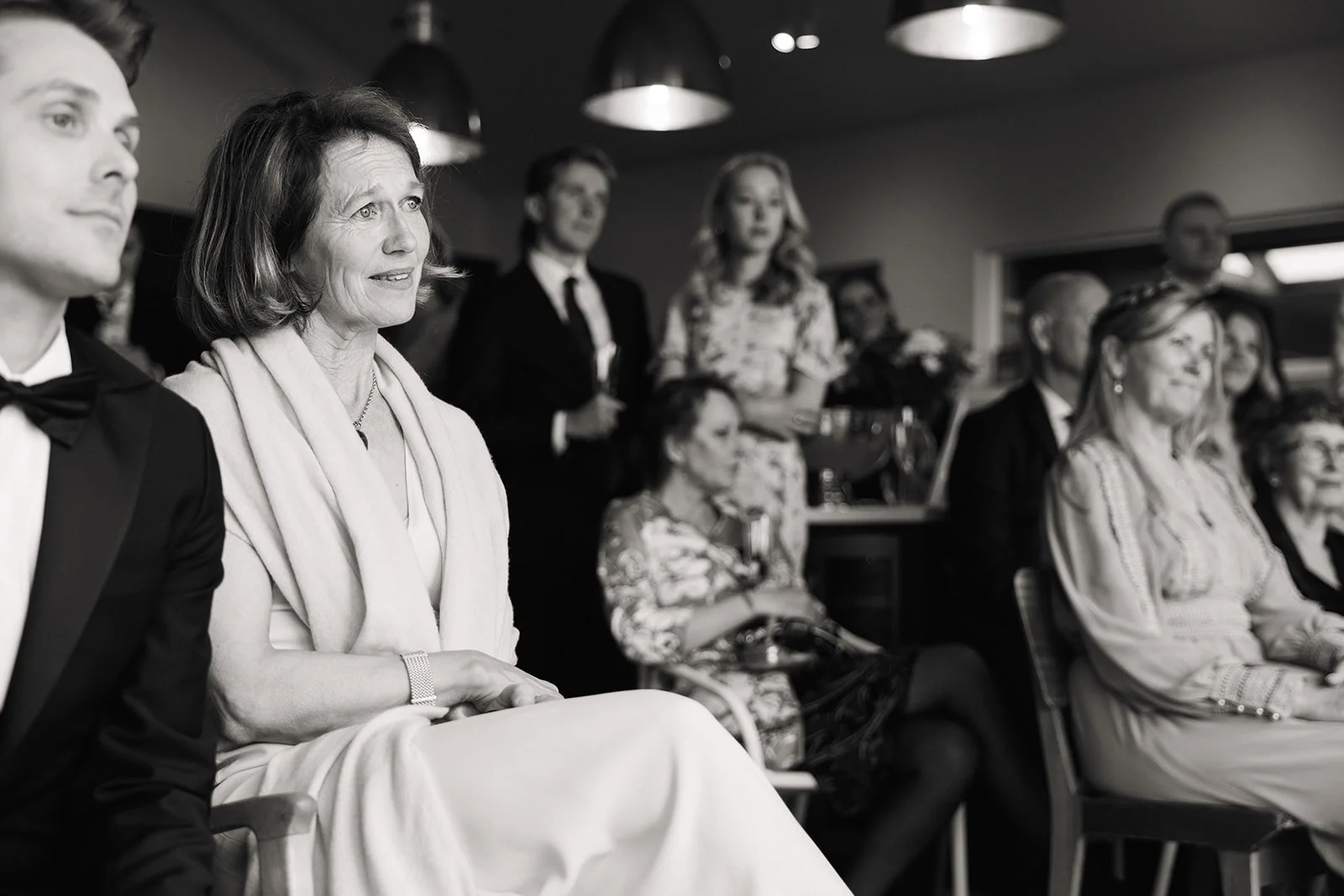 En gruppe mennesker sitter og ser på noe i et rom. De er kledd i formelle klær, og det ser ut til å være en formell ellerseremonielt anledning.