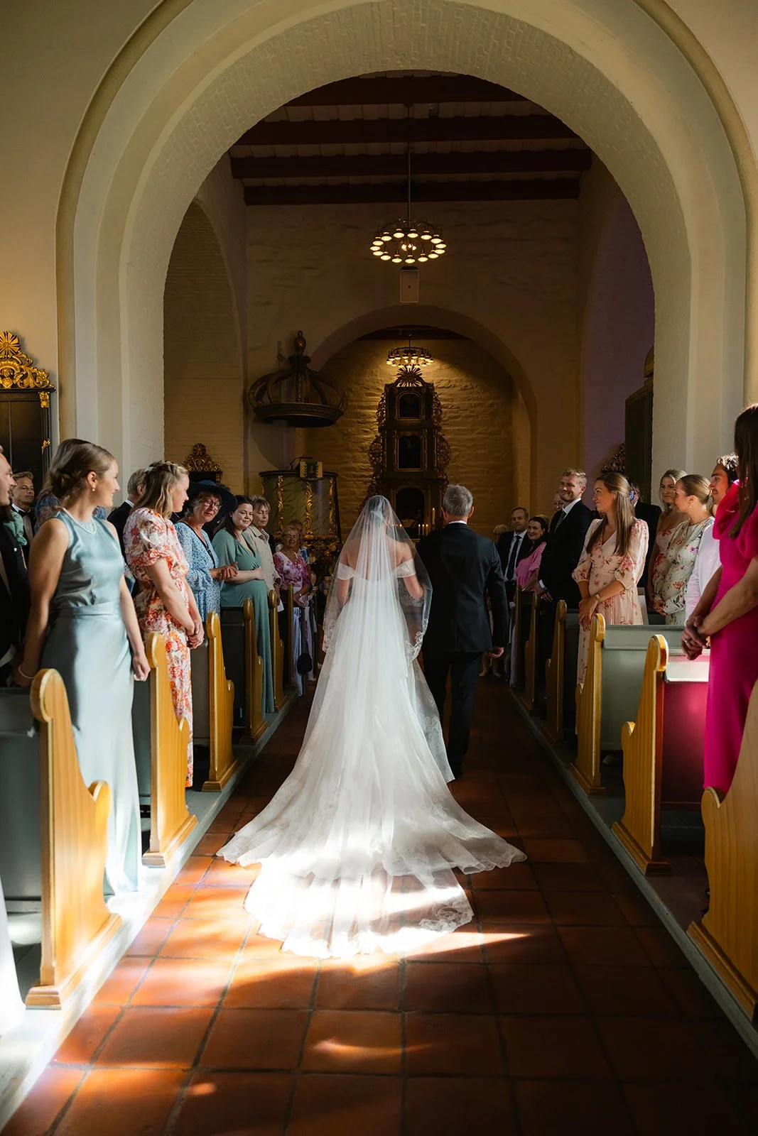 En brud i hvit kjole som går ned midtgangen i en kirke, ledsaget av en person ved hennes side. Kirkens innside er synlig med folk som står på begge sider av midtgangen, og vakker belysning som lyser opp scenen. 