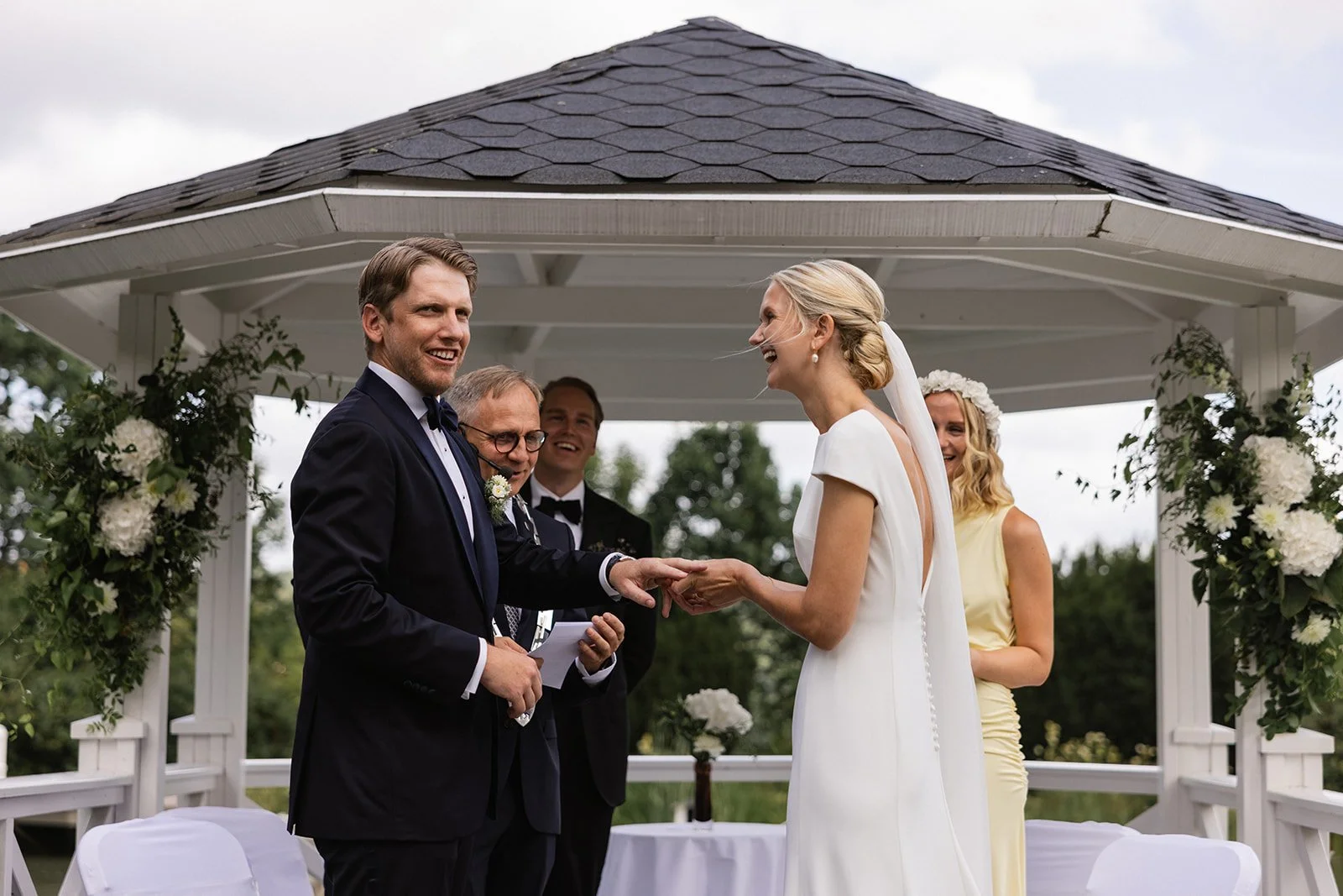 Et brudepar som gifter seg ute i en paviljong, med fire personer som smiler og holder hverandre i hendene, omgitt av blomsterdekorasjoner.
