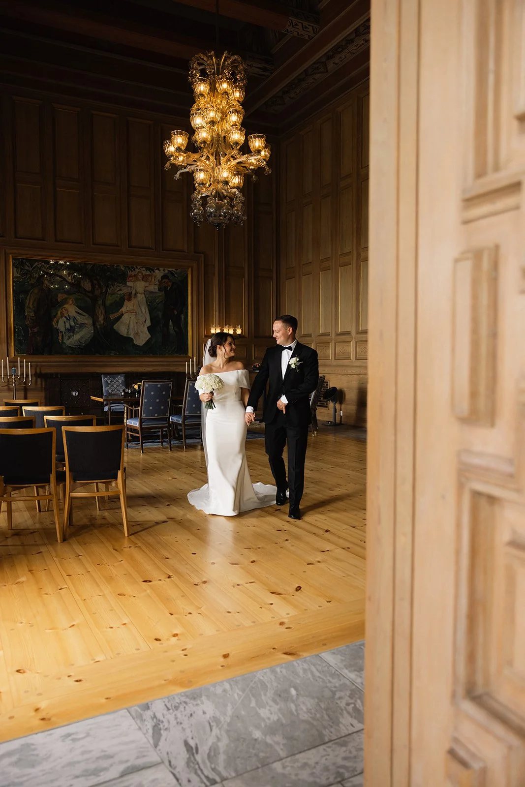 Et ektepar går hånd i hånd inn i en festinngang, omgitt av elegante, klassiske omgivelser med store gylne lysekroner og kunstverk