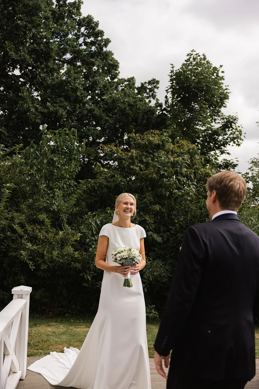 En brud og en brudgom står utenfor, med bruden som smiler og holder en blomsterbukett.