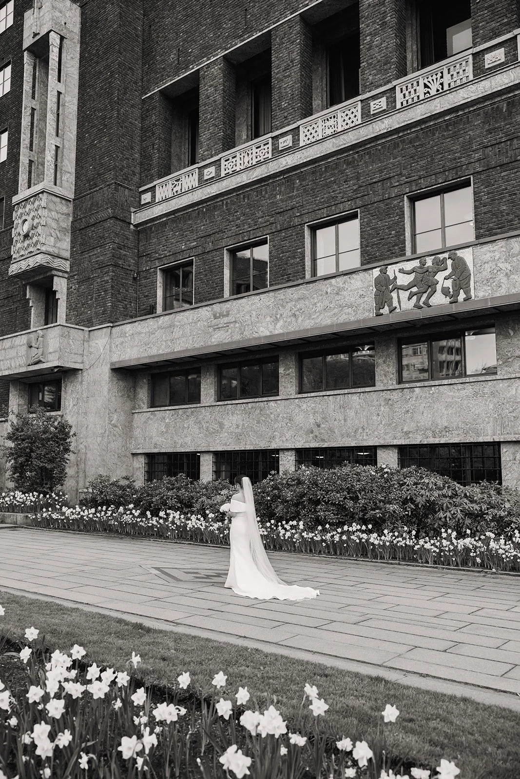 En kvinne i hvit brudekjole står på en gangsti foran en blomstereng med hvite blomster, foran en stor murbygning med mange vinduer og kunstneriske detaljer.
